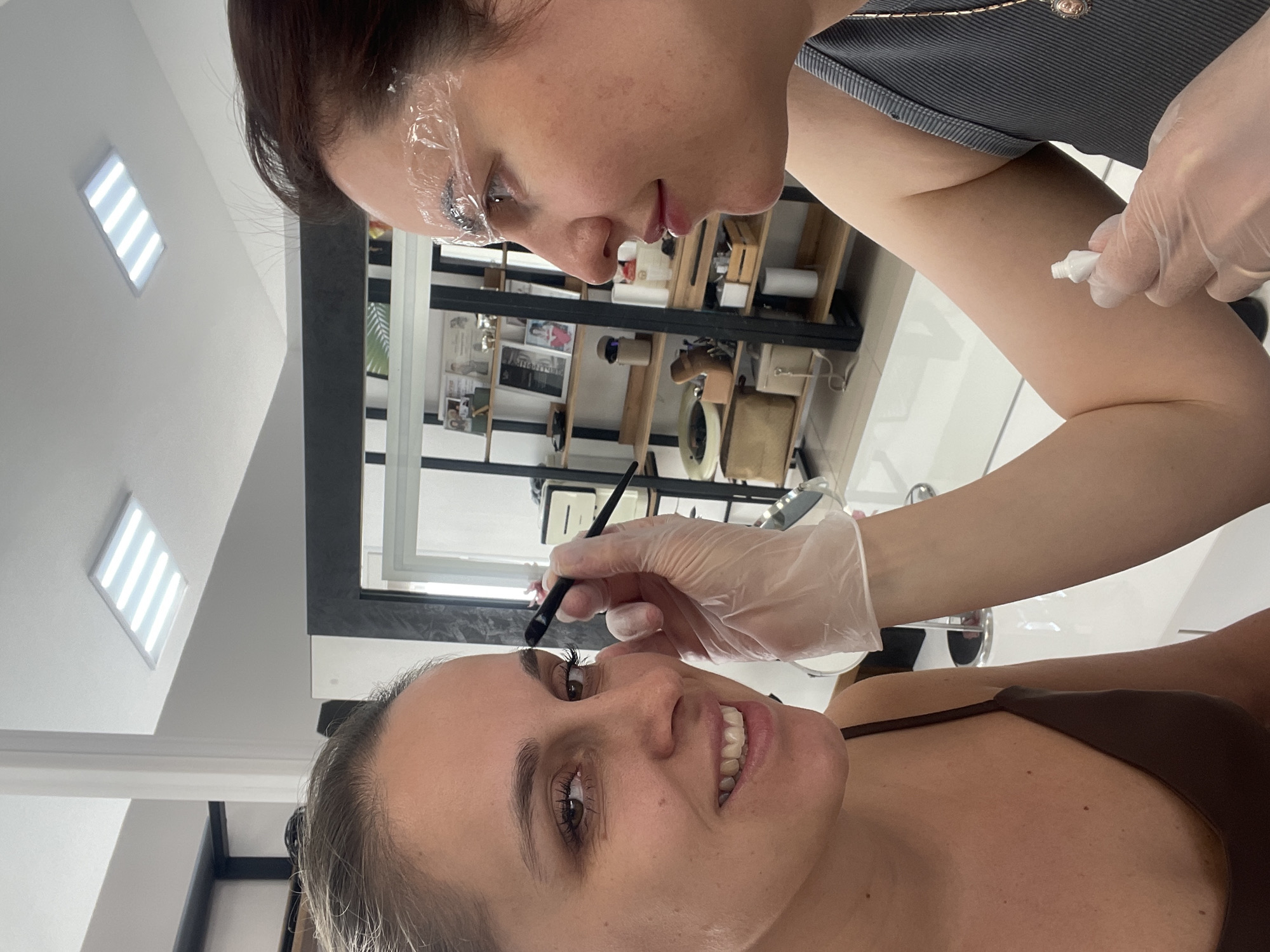 A woman is lying down in a salon in Chaikovsky, Russia, while another woman, wearing gloves, applies something to her eyebrow with a brush. The woman receiving the treatment is smiling. The practitioner has plastic wrap on her eyebrows. There is a shelf with items in the background. The room is well-lit with rectangular ceiling lights. A tube of ointment or cream is visible in the hand of the practitioner.