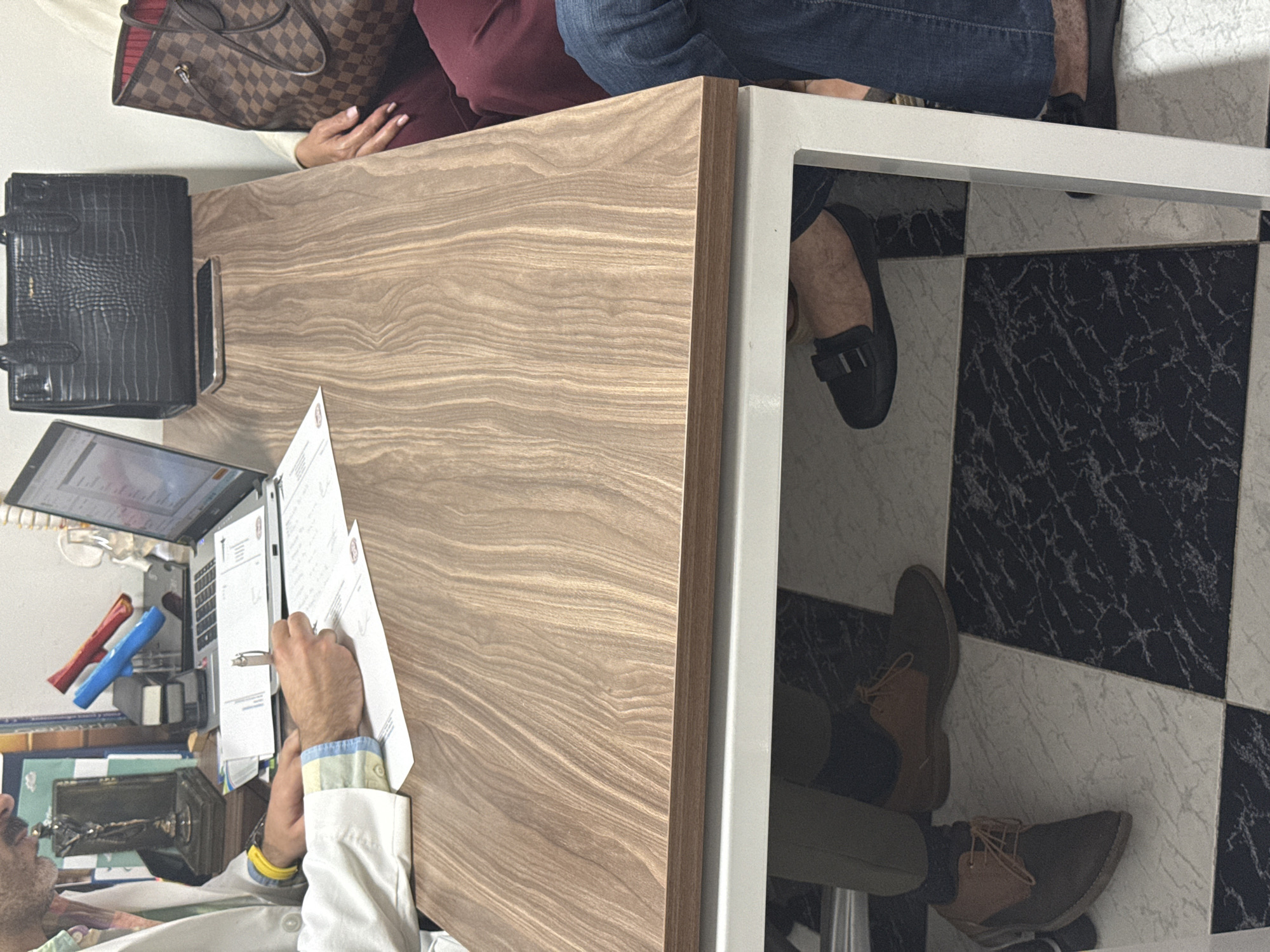 The image depicts a scene in what appears to be a medical office. A man in a white coat is seated at a wooden desk, writing on documents while looking at a laptop. On the desk, there are various items, including pens, a sewing machine, and a black handbag. Two other people are partially visible: one seated on the desk wearing jeans and another sitting under the desk wearing brown shoes. The office has a black and white checkered floor and is furnished with a table that has a white metal frame. The location is speculated to be in Puebla, Mexico, based on elements or inferences from the broader context that may be beyond this image alone.