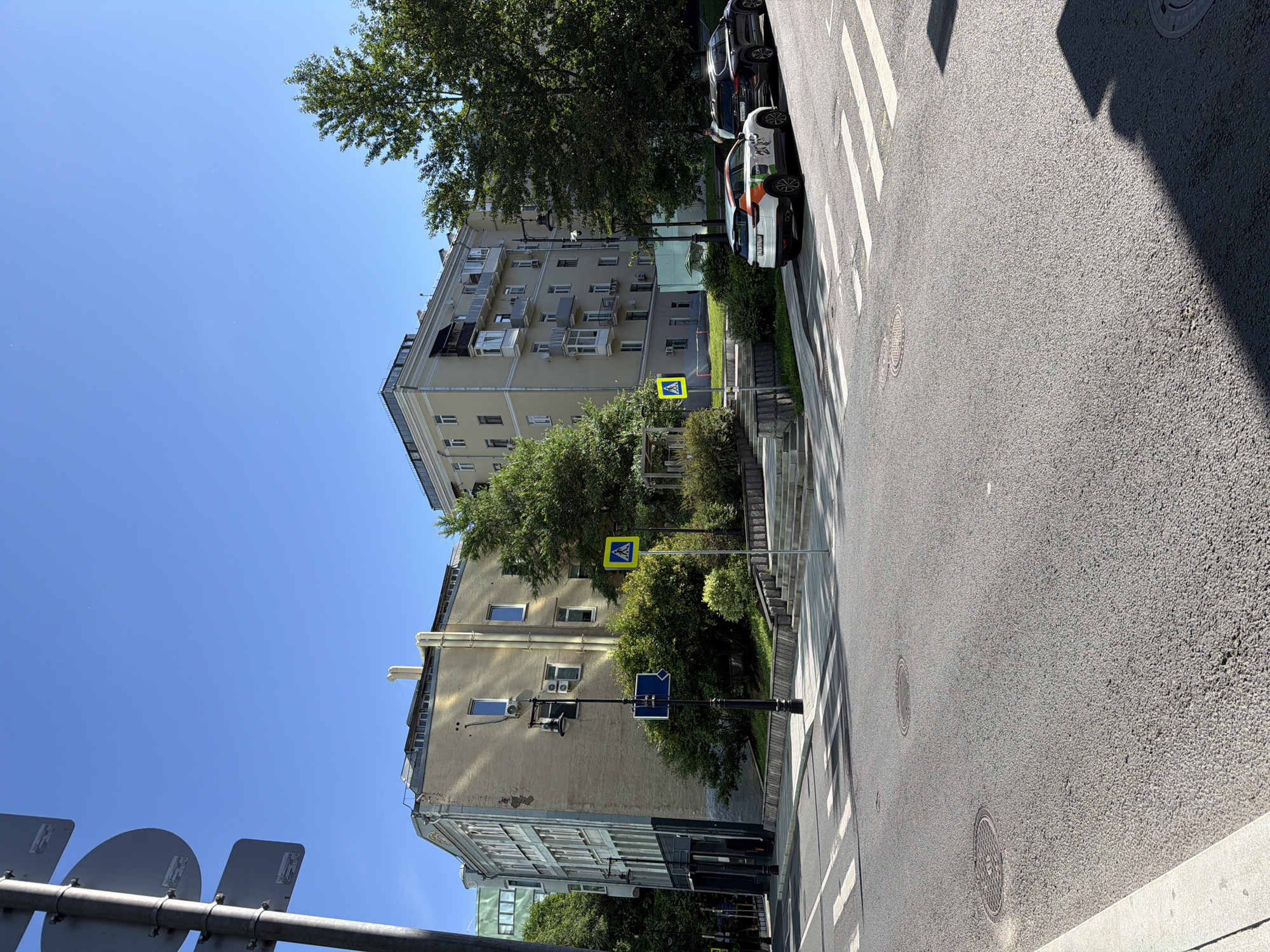 The image shows a street in Moscow, Russia, with a clear blue sky overhead. In the foreground, there is a paved road with a pedestrian crossing. Two cars are parked on the right side of the road: a white electric car with colorful graphics and a black SUV. Behind the cars, a light-colored residential building with multiple balconies and windows stands tall. Adjacent to the building, lush green trees and bushes add to the urban landscape. To the left, another residential building, taller and with a light green facade, is partially visible. A street lamp with a camera attached and a blue road sign are also present on the left side of the image. Two yellow pedestrian crossing signs are visible, one on the left and one on the right side of the road. The scene depicts a typical day in a Moscow neighborhood, with a mix of architecture, nature, and urban infrastructure.