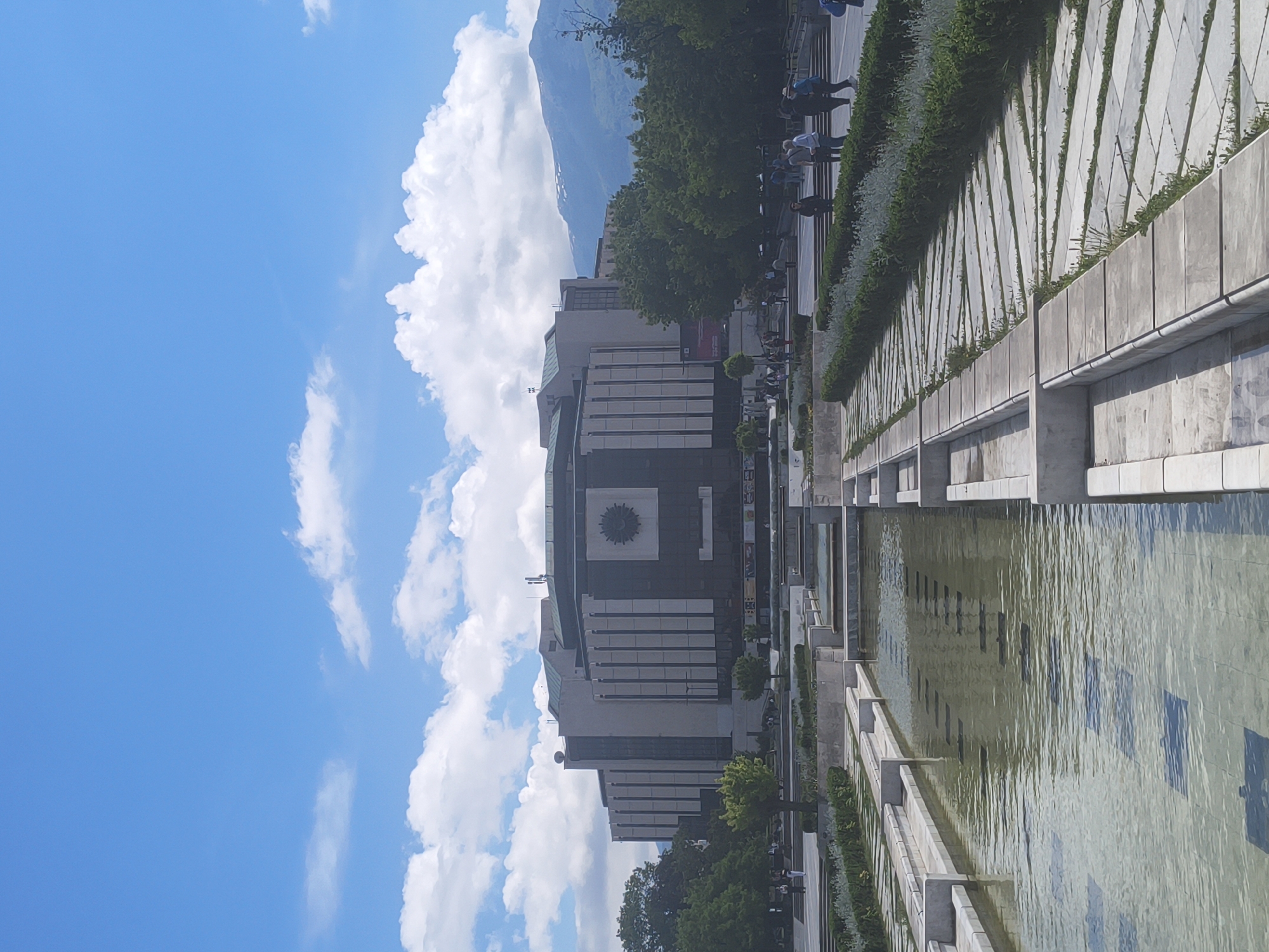 The image shows the National Palace of Culture in Sofia, Bulgaria, viewed from a ground-level perspective across a long, rectangular reflecting pool. The building, composed of multiple stacked horizontal layers, features a decorative central element. The pool's edges are defined by concrete structures with planted greenery. A sidewalk runs alongside the greenery, with people walking along it. The sky is bright blue with scattered white cumulus clouds. A mountain is visible in the background, behind the palace.