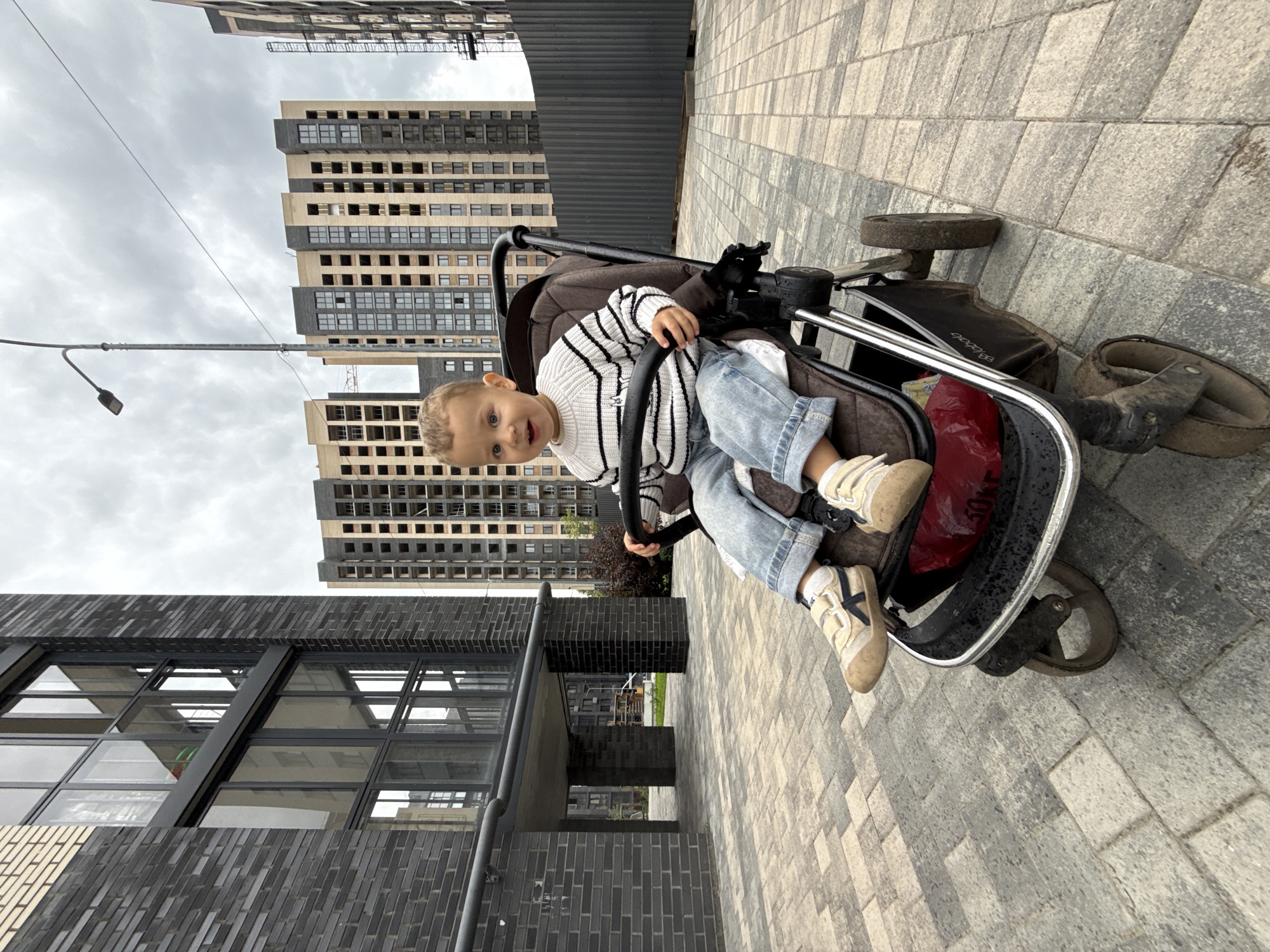 An infant wearing a striped sweater and light blue jeans sits in a stroller on a paved area in front of a modern brick building with large windows. The background features tall residential buildings under construction and a grey, cloudy sky. The scene is likely located in Rusanovo, Russia.