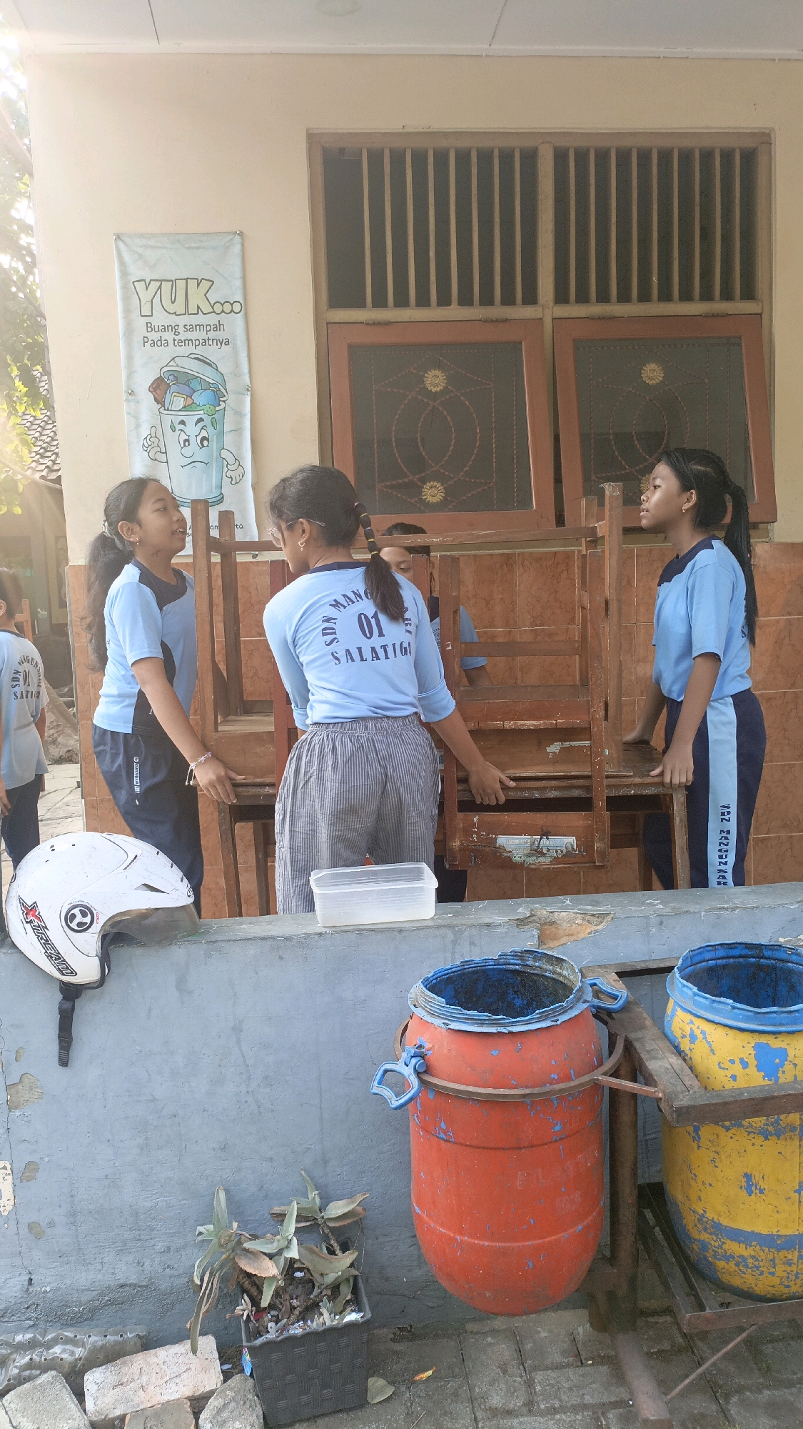 In Salatiga, Indonesia, three young girls wearing blue and navy school uniforms are moving wooden chairs. The girl on the left, with dark hair in a ponytail, faces the center and speaks with the girl beside her. The girl in the middle, facing away from the camera, has her hair in a ponytail and wears glasses. The girl on the right, also with dark hair in a ponytail, faces to her right. They are in an outdoor area with a light-colored building and a window with vertical bars in the background. A banner with the text "YUK... Buang sampah Pada tempatnya" and an illustration of a trash can is visible on the wall. In the foreground, a white helmet rests on a gray ledge. Two large plastic barrels, one orange and one yellow, are positioned near the girls. A potted plant with succulent-like leaves sits next to the barrels. The ground appears to be paved with bricks.
