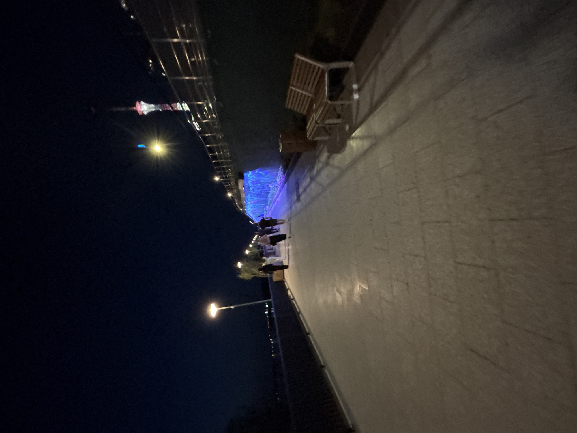 This is a nighttime scene along a waterfront promenade in Perm, Russia. A paved walkway stretches into the distance, illuminated by streetlights. A wooden bench is visible beside the walkway. People are walking along the promenade towards the center of the image. A distinctive tower with illuminated elements is seen in the background. The riverbank is reinforced with a concrete structure. The sky is dark, indicating the time of day.