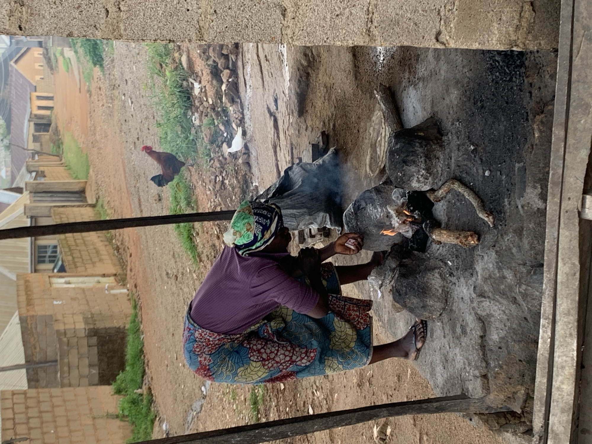 A woman, wearing a purple shirt and a colorful patterned skirt and headscarf, is bent over a makeshift fire pit on a concrete platform. She appears to be tending to the fire, with smoke rising from it. Several pieces of wood are scattered around the fire pit, and some dark grey rocks are used to support the cooking area. Behind her, a rooster stands on the dusty ground amidst sparse green grass and scattered rocks. In the background, there are buildings constructed from brick and cement, with corrugated metal roofs, typical of residential areas in Jos, Nigeria. The scene depicts an outdoor cooking activity in a semi-urban or rural setting.