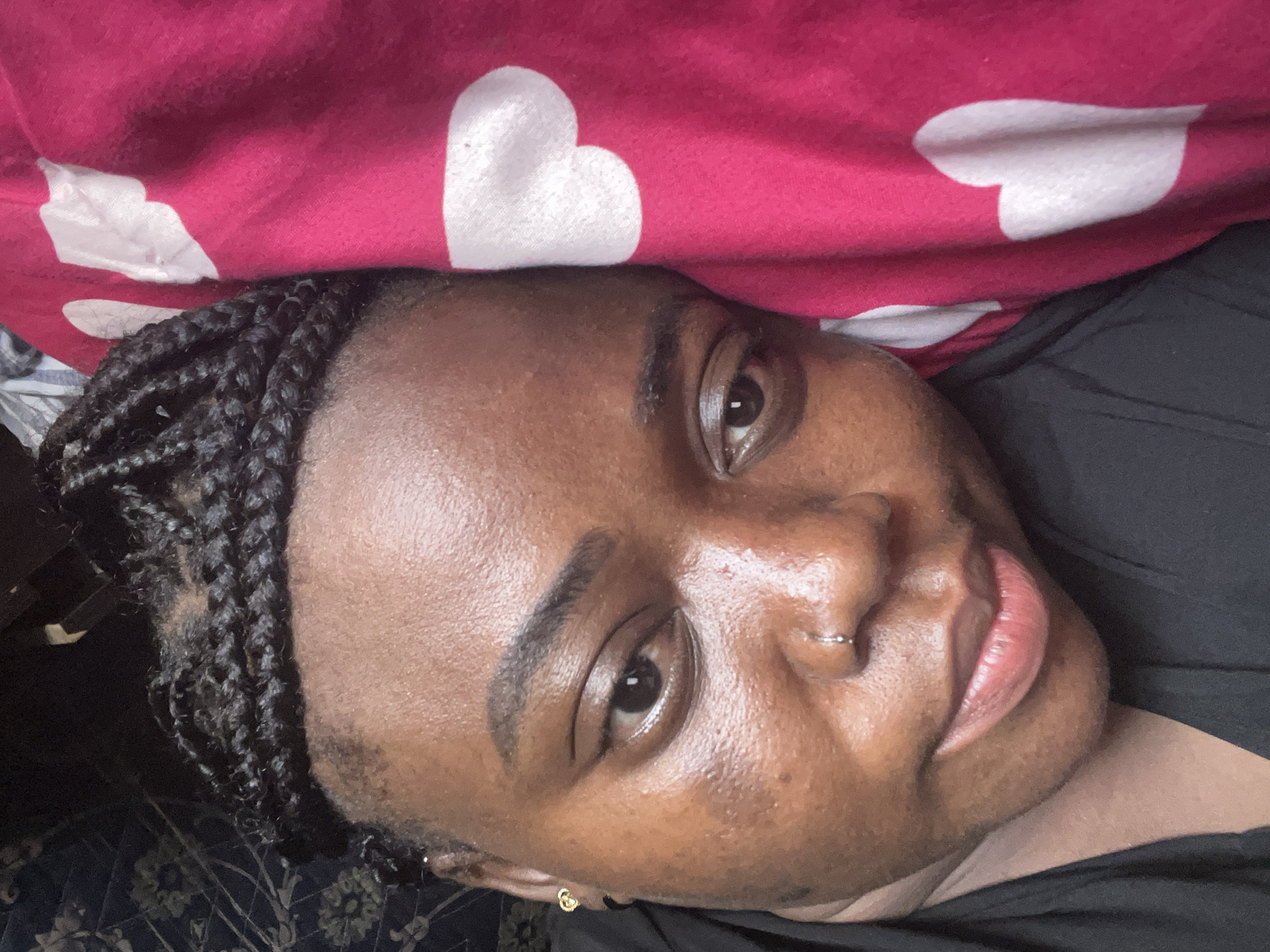 A close-up, portrait-style image features a dark-skinned individual, likely a woman, lying down and looking directly towards the camera. Her head is angled, with braided hair styled in cornrows visible on the left side of the frame. She wears a dark, possibly black, piece of clothing, with a small golden, heart-shaped earring visible in her left ear.
Her face is well-lit, revealing dark eyes, shaped eyebrows, and full lips with a natural hue. A small silver hoop piercing adorns her right nostril. The individual has a relaxed and soft expression, with a subtle hint of a smile.
In the background, a vibrant pink fabric, possibly a blanket or pillow, is visible, decorated with several white heart patterns. One large, clear white heart is prominent above her head. The setting appears to be indoors and casual, suggesting a moment of rest or relaxation. This scene takes place in Abuja, Nigeria. No visible text is present in the image.