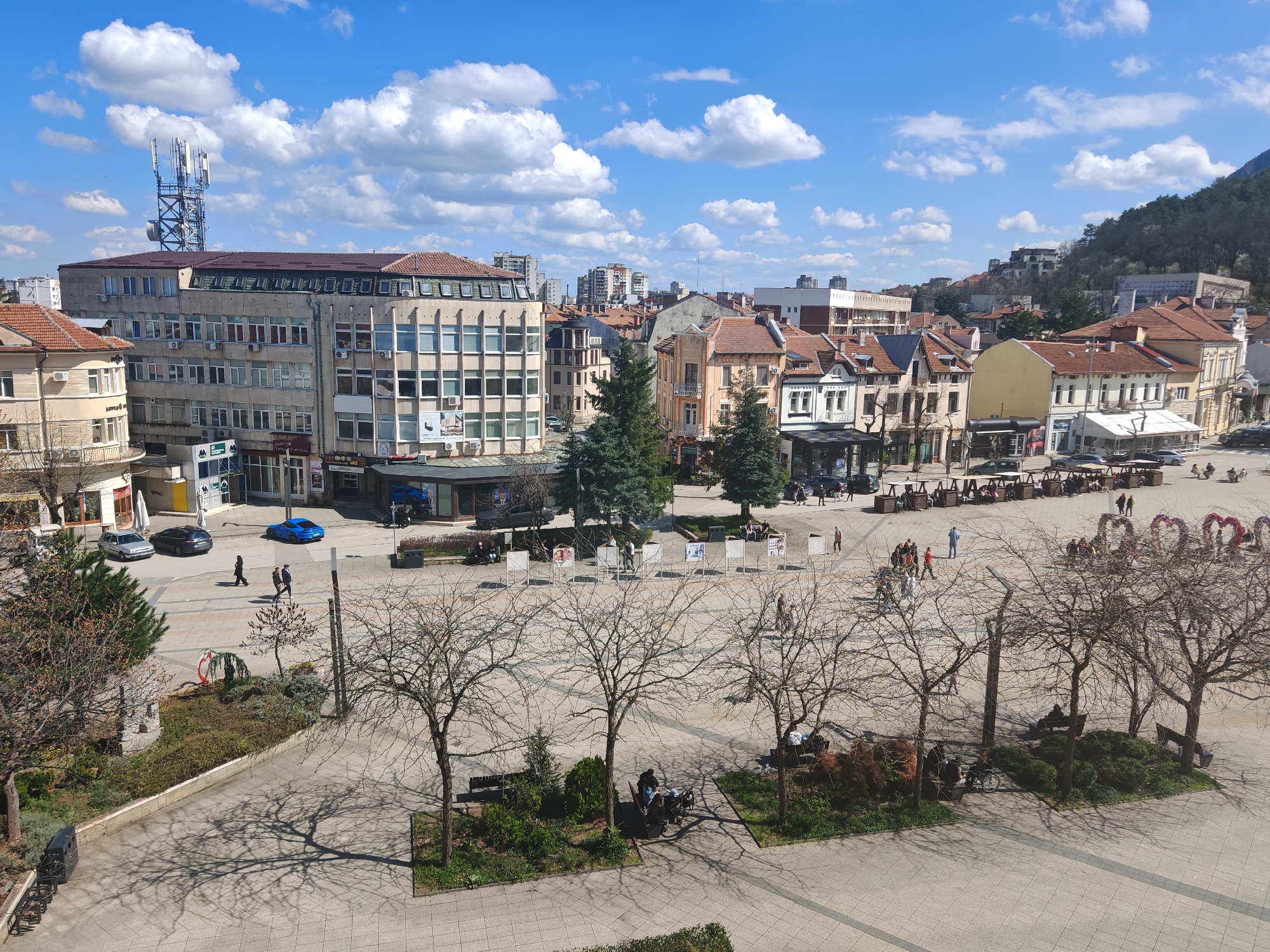 The image shows an elevated daytime view of a large public square, likely a central plaza, in the city of Vratsa, Bulgaria. The square is paved with light-colored square tiles and features several bare-branched trees, small landscaped islands with benches, and walking paths. People are seen strolling and sitting on the benches.
In the background, there's a mix of architectural styles, including older buildings with red tile roofs and more modern, concrete structures. A telecommunications tower rises above the skyline.
The weather appears sunny with scattered clouds. Cars are parked on the street adjacent to the square. The location can be identified by its unique urban layout, architectural design, and the surrounding landscape.