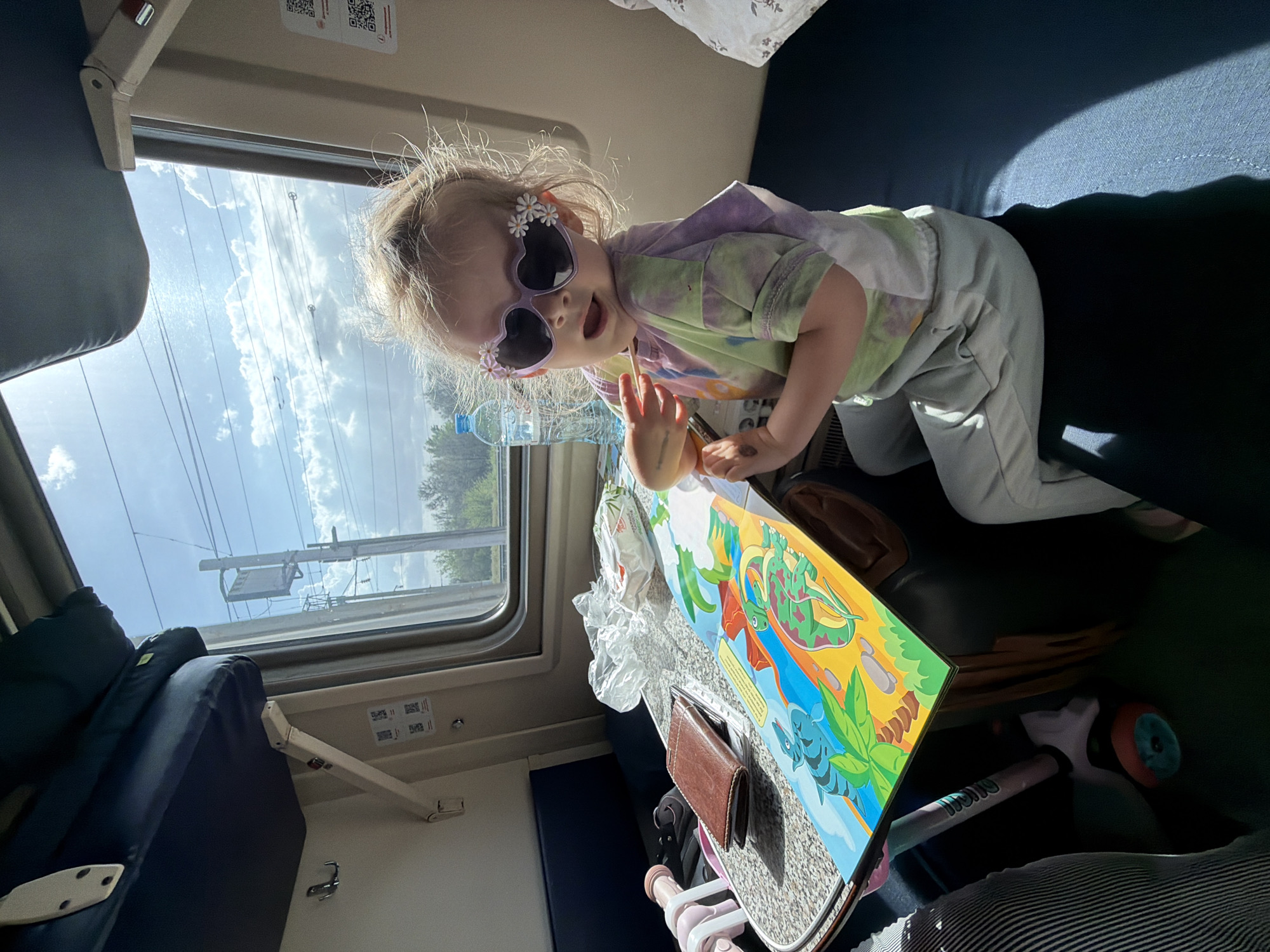 A photograph shows a child wearing heart-shaped sunglasses with flower embellishments, reclining on a blue seat and reaching for a book. The book, which appears to be resting on a small table, features brightly colored cartoon dinosaurs and other illustrations. A brown wallet also sits on the table. A water bottle and white packaging are next to the book. A scooter is next to the table. Outside the window, the sky is visible, and trees can be seen in the distance. The child is wearing a tie-dye shirt, light-colored pants, and a hoodie. The photo was taken in the city of Petrov Val, Russia.