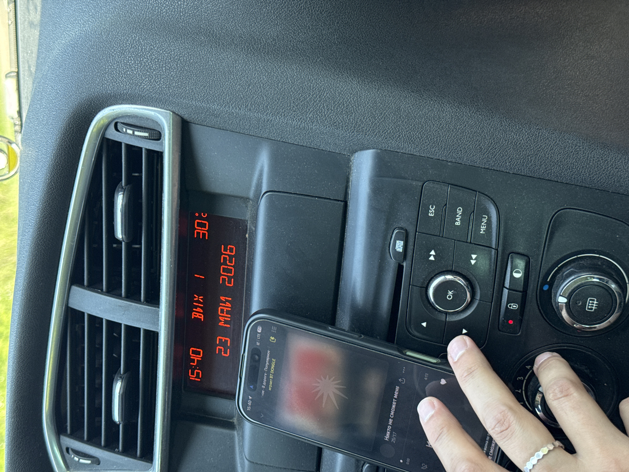The image shows the dashboard of a vehicle. A smartphone rests on the dashboard, displaying audio information. The time shown on the phone and the car's display reads 15:40. The car's display also shows the date as 23 May 2026 and a temperature of 30°C. A hand is shown operating the vehicle's controls. The environment implies the interior of a car, but without further context, the specific location in Shaberdino, Russia, cannot be determined.