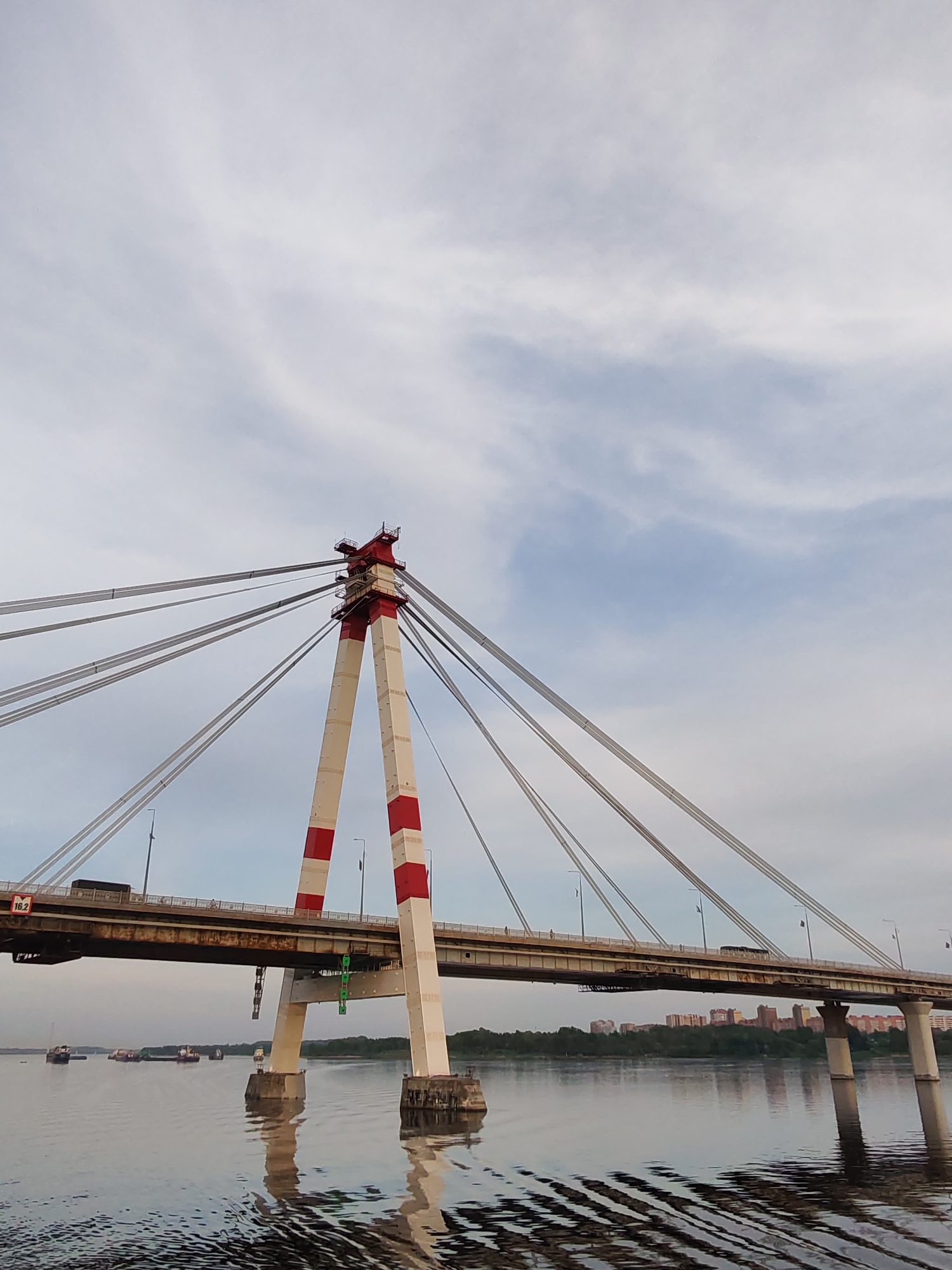 A low-angle shot shows a bridge that stretches across a body of water. The bridge's main support structure is a tall, white pylon adorned with broad red stripes on its upper and lower sections. Cables extend from the top of the pylon to the bridge's deck, creating a web-like pattern against the sky.
The bridge's deck is a gray, roadway surface, with a few vehicles visible. Streetlights line the bridge, adding to the structural detail.
Below the bridge, the water reflects the bridge's structure, including the pylon and its supporting pillars. Gentle ripples disturb the water's surface, creating a textured effect.
In the distance, a line of trees forms a horizon against which low-rise buildings can be seen. The sky above is overcast with a mix of white and gray clouds.