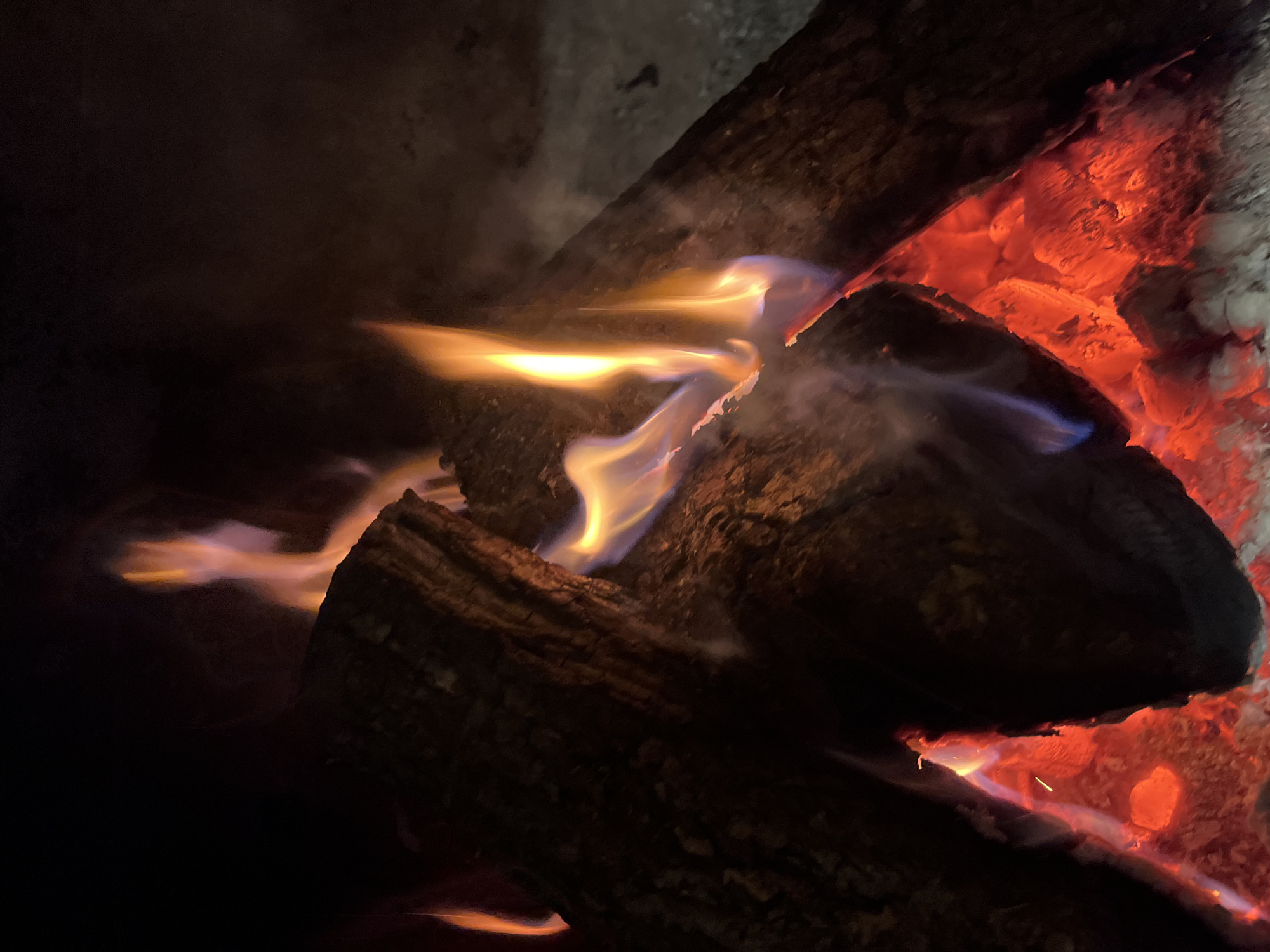 A close-up shot depicts a campfire at night. Multiple charred logs are arranged within the fire. Flames are visible, displaying hues of yellow, orange, and blue. Some embers appear as a glowing red. The background is dark. The scene appears to be outside but the image does not provide any other information about location.