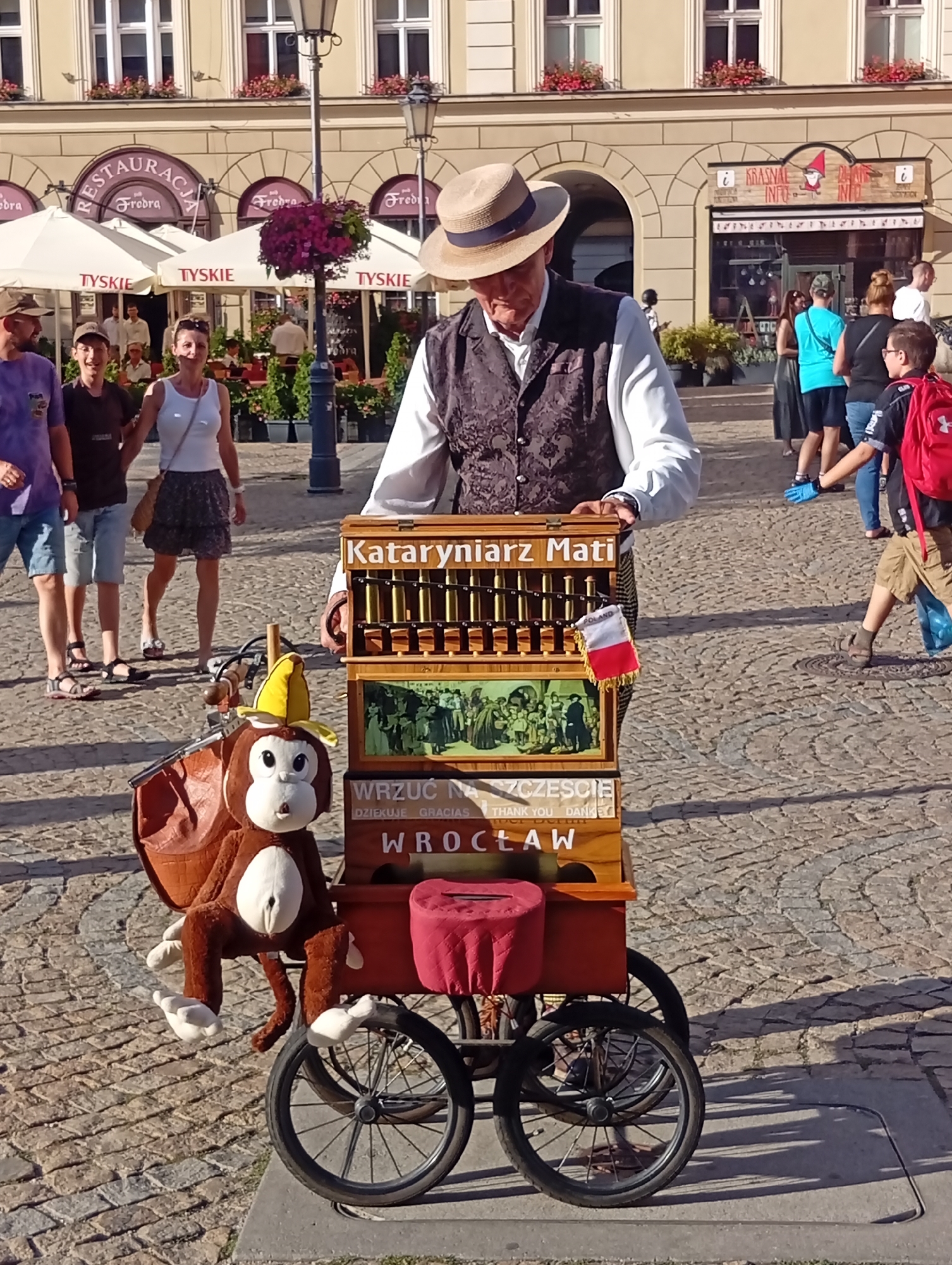 The image depicts a street scene in Wrocław, Poland, on a sunny day. The foreground is dominated by a street organ, a wooden music box mounted on a cart with two large wheels. A stuffed monkey wearing a yellow hat is attached to the front of the cart. The street organ has several levels, with a decorated front panel featuring what appears to be a painted scene and text in Polish. Above this, a row of organ pipes is visible, and a small Polish flag is attached to the side. A man in a straw hat, white shirt, and a dark, patterned vest stands behind the organ, operating it. In the background, several people are walking, including a family in casual clothing. Buildings with shops and restaurants are visible behind them, with awnings providing shade. The ground is paved with cobblestones.