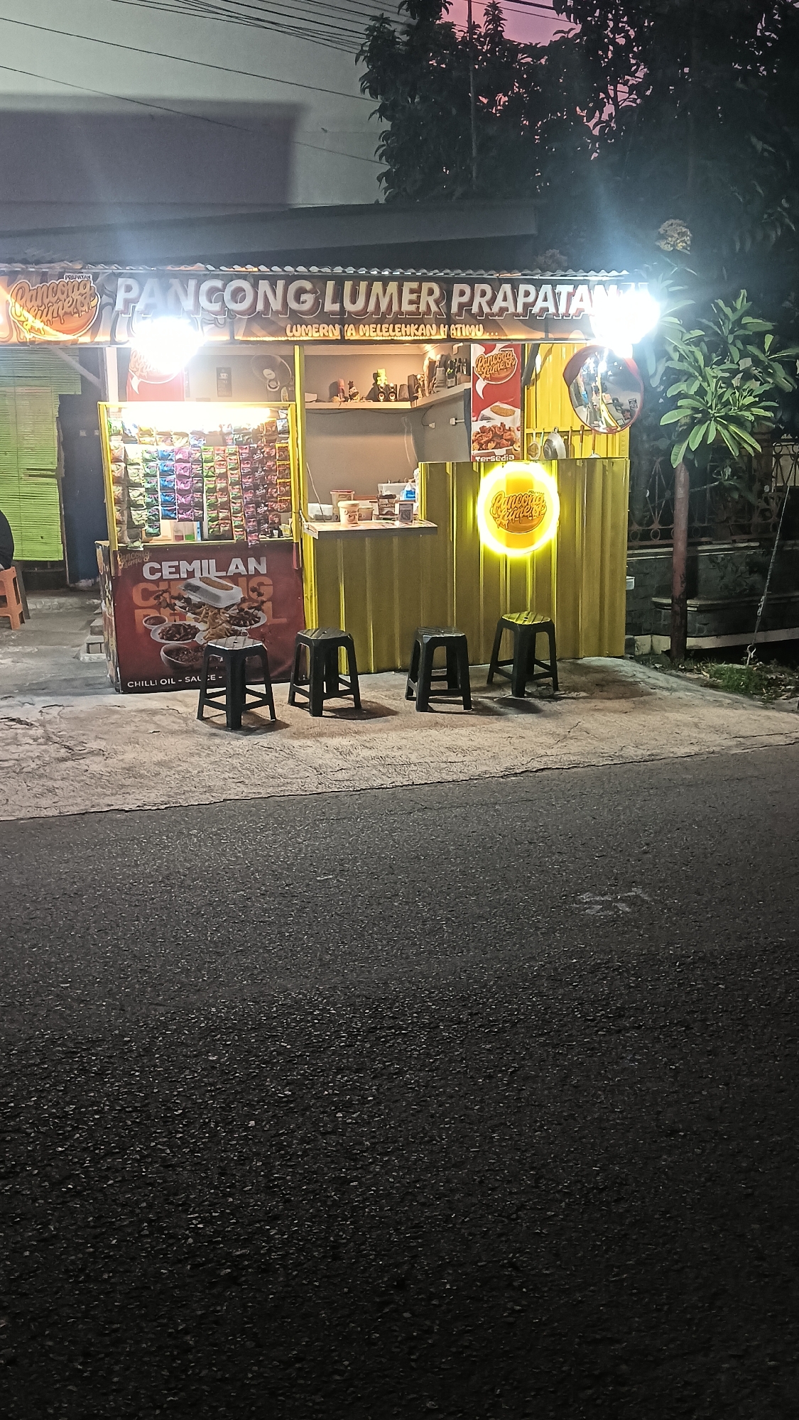 A street food stall named "Pancong Lumer Prapatans" is captured at night on a street in Sragen Tengah, Indonesia. The stall features a bright yellow facade with a lit sign. Various snacks are displayed on shelves behind a counter. A large red sign advertising "Cemilan Chilli Oil Sauce" is prominently displayed. Four black stools are placed in front of the stall. The scene is illuminated by the stall's lights and the fading evening sky.