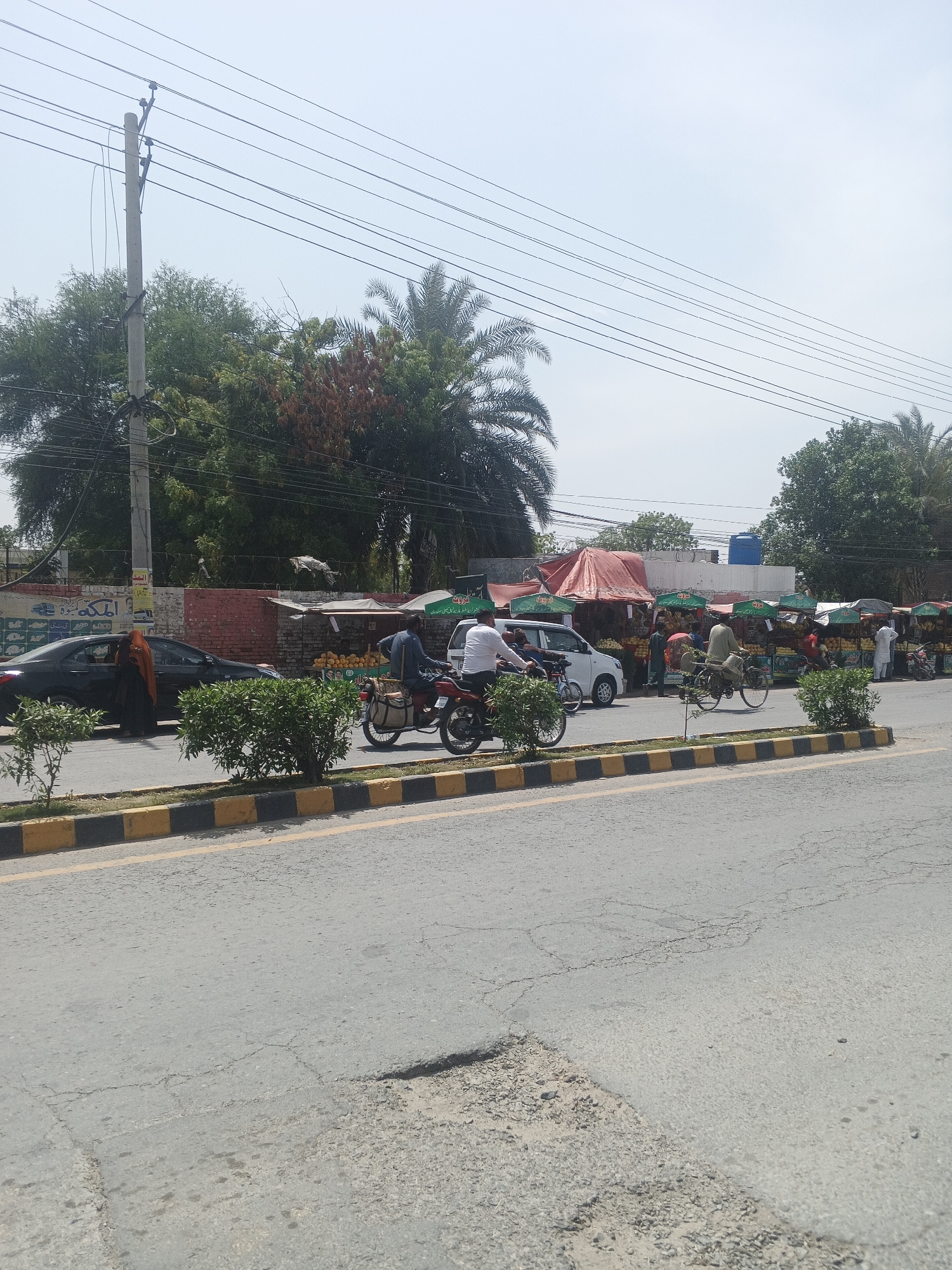This image captures a vibrant street scene in Samundri, Pakistan, depicting daily life and commerce under a clear, sunny sky.
In the foreground, a wide asphalt road, showing signs of wear with visible cracks, stretches across the frame. It is bordered by a yellow and black painted curb that forms a divider, dotted with small green bushes. On the road, a man rides a red motorcycle, carrying a large canvas bag, while further down, another man cycles on a bicycle.
To the left, a black sedan is either parked or stopped, and a person, possibly a woman, clad in an orange or brown head covering, is seen partially exiting or entering the vehicle. Along the side of the road, a series of bustling makeshift market stalls are set up beneath red and green awnings, displaying piles of fresh fruits, likely mangoes. Several individuals, appearing to be vendors and customers, are scattered around these stalls. A white van is also visible near these market stands.
The background features a variety of lush green trees, including distinctive palm trees, with some foliage showing reddish-brown hues. A prominent concrete utility pole on the left supports a dense network of overhead power lines and wires that crisscross the bright, pale blue sky. Behind the black car, a brick wall is visible, adorned with various signs featuring Urdu script, including the word "الحَمْدُ" (Alhamdu), indicating a commercial or public message. Further down, a white building with a blue water tank on its roof can be seen amidst the trees.
The scene conveys a typical, active daytime environment in a South Asian town, characterized by street vendors, diverse modes of transport, and the ubiquitous presence of overhead electrical infrastructure.