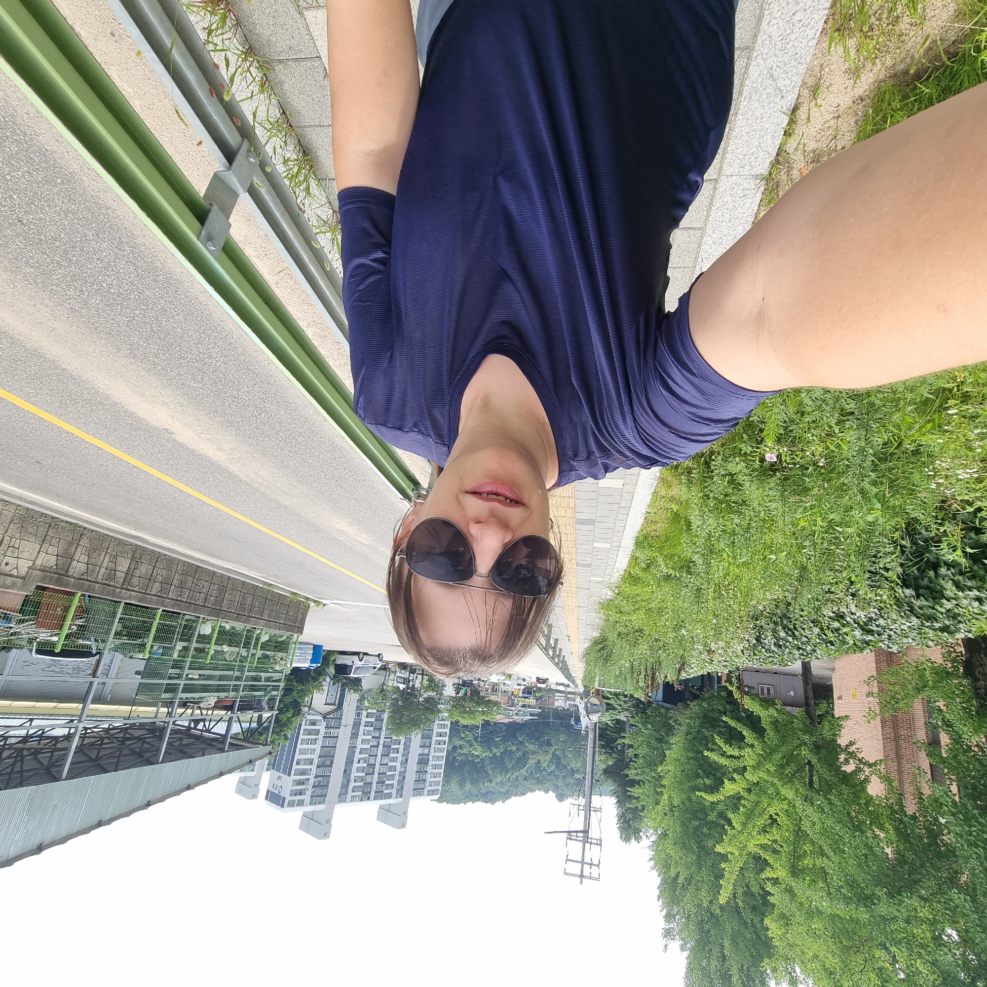 A selfie is taken by a woman standing on a sidewalk in Dunnae-myeon, South Korea, facing the camera. She is wearing a dark blue t-shirt and sunglasses. To her right is a road with a yellow line, a green guardrail, and a concrete wall. To her left is a sidewalk, a grassy area with some small flowers, and a brick building. In the background, there are trees, a utility pole, a cluster of apartment buildings, and a covered outdoor structure. The sky is overcast.