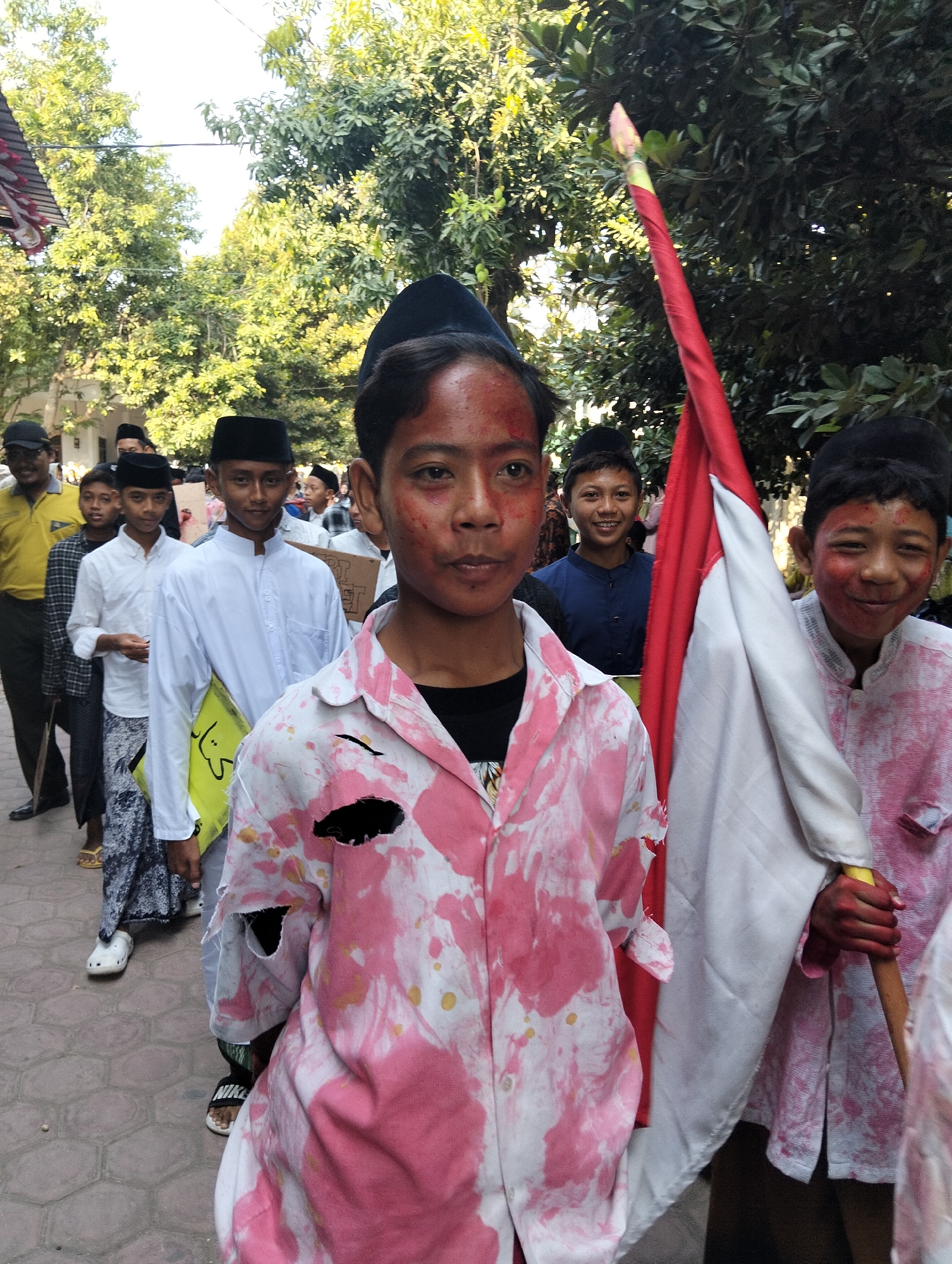 In Kranji, Indonesia, a group of young boys, some with red paint or makeup smeared on their faces, stand in a line. The boy in the foreground wears a tattered pink and white shirt, and his face is covered in red marks. He is holding a flag pole. To his right, another boy in a similar pink and white shirt also has red markings on his face and is holding a red and white flag. Behind them, other boys are visible, some wearing white shirts and black caps. The scene appears to be outdoors, with trees and a building in the background. The ground is paved with hexagonal stones.