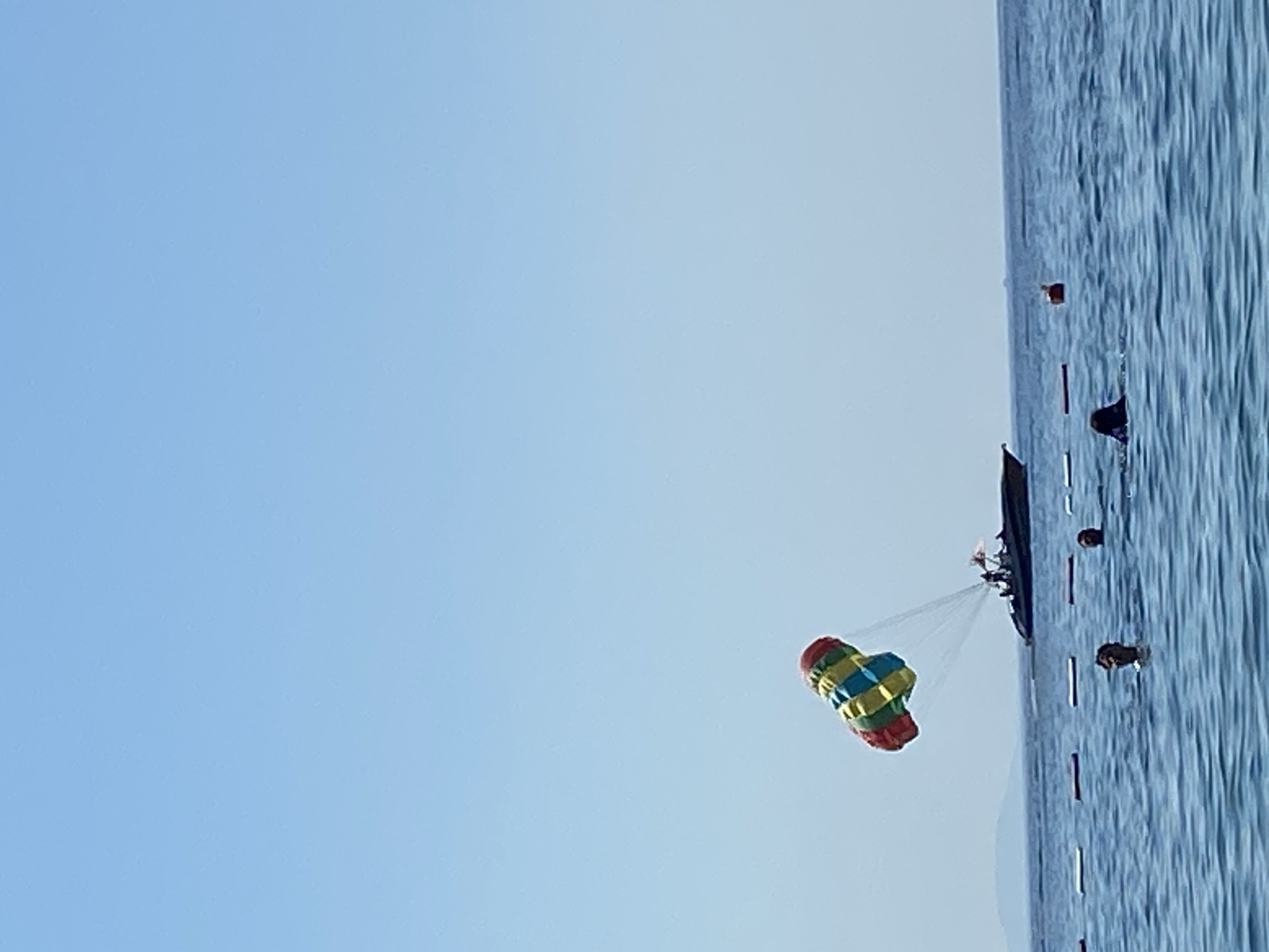 The image depicts a parasailing activity taking place over the sea near Budva, Montenegro. The scene is dominated by a vast expanse of clear blue sky, suggesting fair weather, likely during the daytime given the bright light. The water, a deep blue with visible ripples, occupies the right side of the frame, with a boat visible just above the waterline. Attached to the boat by tow ropes is a colorful parasail, adorned with red, yellow, and blue stripes. A person or people are suspended beneath the parasail, though they are not clearly discernible. The boat appears to be a functional vessel, with some dark objects or flags attached to its stern. The overall impression is one of leisure and outdoor recreation, typical of a coastal resort. There is no visible text in the image.