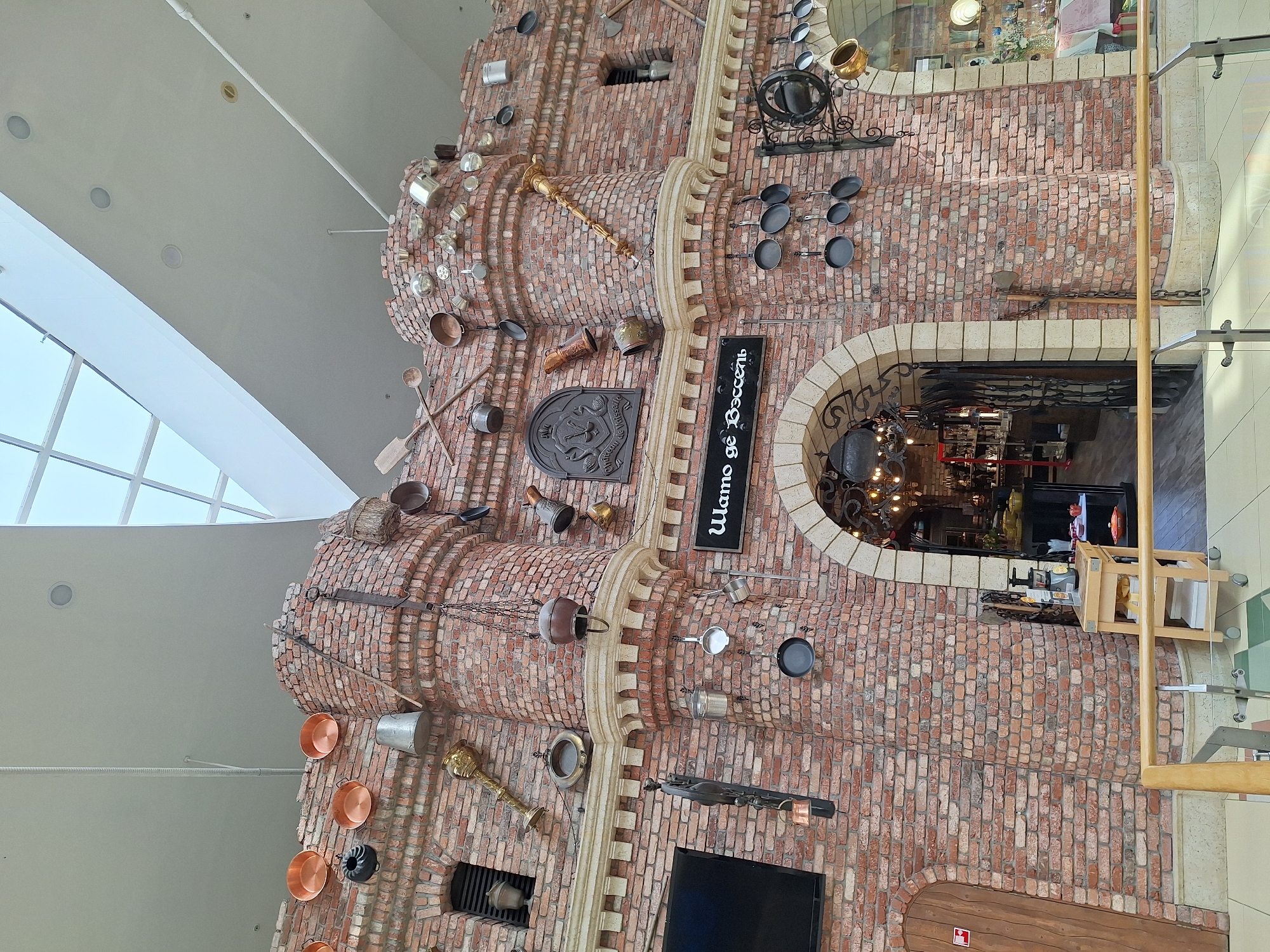 The image displays a multi-story interior facade of a building, reminiscent of a medieval castle or fortress wall, elaborately decorated with numerous kitchen-related objects. This rustic brick structure, located in what appears to be a modern indoor commercial space, likely in Moscow, Russia, features various shades of red and brown bricks, forming arched windows and decorative turrets.
The brickwork is extensively adorned with a wide array of metal and copper pots, pans, ladles, spatulas, colanders, and other vintage kitchen utensils and cookware of different sizes and designs. These objects are hung or attached to the brick surface, creating a vibrant, themed display. Notable decorative elements include a large gold-colored pestle or mace-like object, a dark crest or shield design, and decorative stone-like columns integrated into the brick.
An arched entrance on the right side of the structure leads into a dimly lit interior, revealing chandeliers and what appears to be a restaurant or shop setting. A tall, black vertical sign with white text is prominently displayed on the brick wall. The text reads "Шато де Восель" (Château de Vaucelles), suggesting a French-themed establishment.
The ceiling above the structure is high and light-colored, featuring recessed lighting and large skylights with white frames that allow natural light to illuminate the scene, indicating daytime. A modern glass and wooden railing is visible on the far right, suggesting an upper level or balcony overlooking the decorated wall. No people are visible, and the scene is static, showcasing the architectural and decorative elements of the establishment.