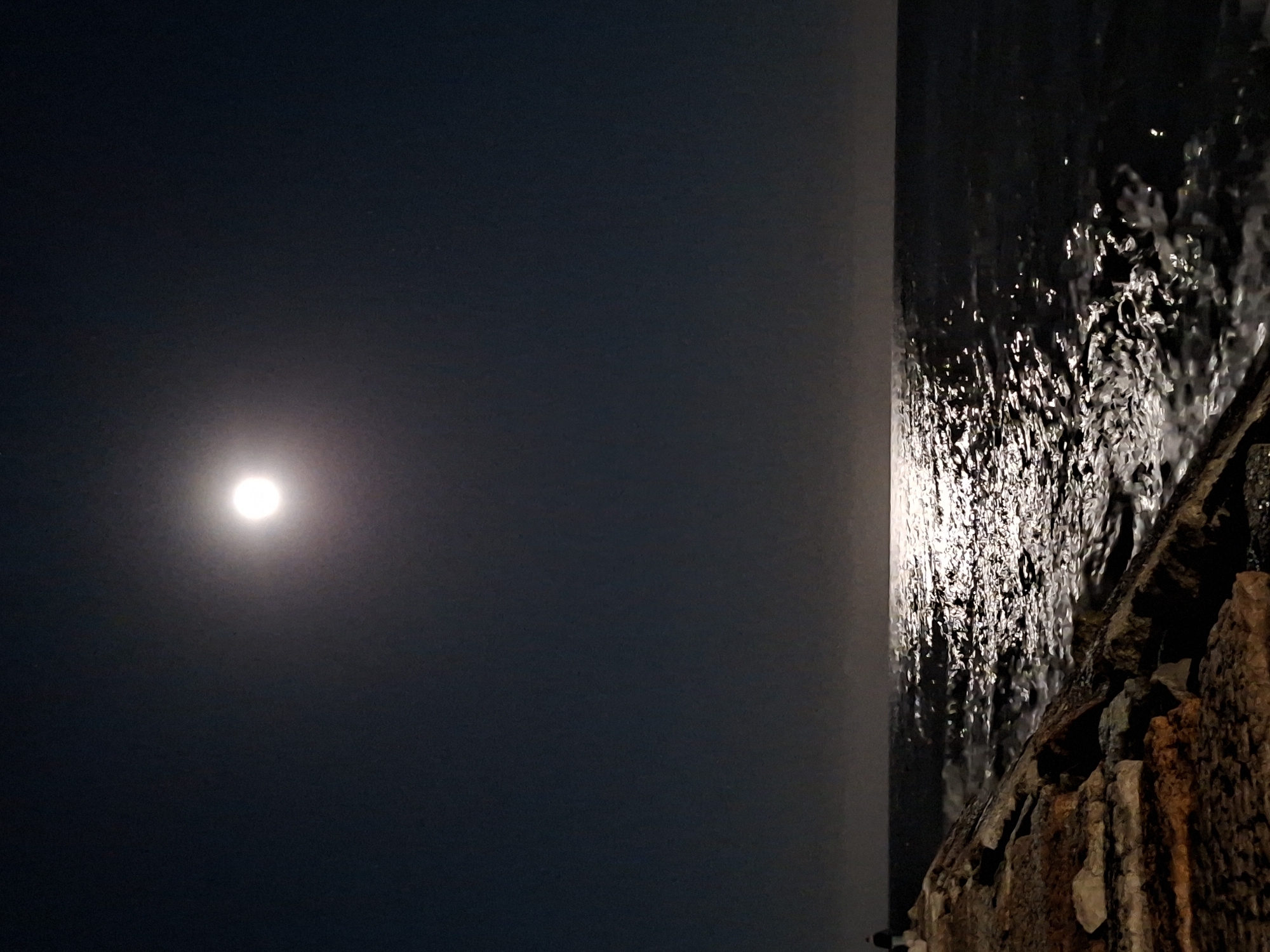 The image depicts a nighttime scene at the sea, likely in Caorle, Italy, characterized by the bright illumination of a full moon. The moon is positioned in the upper center of the dark sky, casting a strong, shimmering reflection on the water's surface. In the foreground, a rocky jetty or seawall extends from the lower left corner, with rough, uneven stones of varying shades of brown and gray. The sea is dark and choppy, with waves breaking and creating highlights from the moonlight. A solitary figure, silhouetted and small in scale, stands on the jetty near the left edge of the frame, facing towards the sea. The horizon is faintly visible as a blurred line between the sea and the sky. The overall atmosphere is serene and nocturnal, emphasizing the natural light of the moon over the ocean.