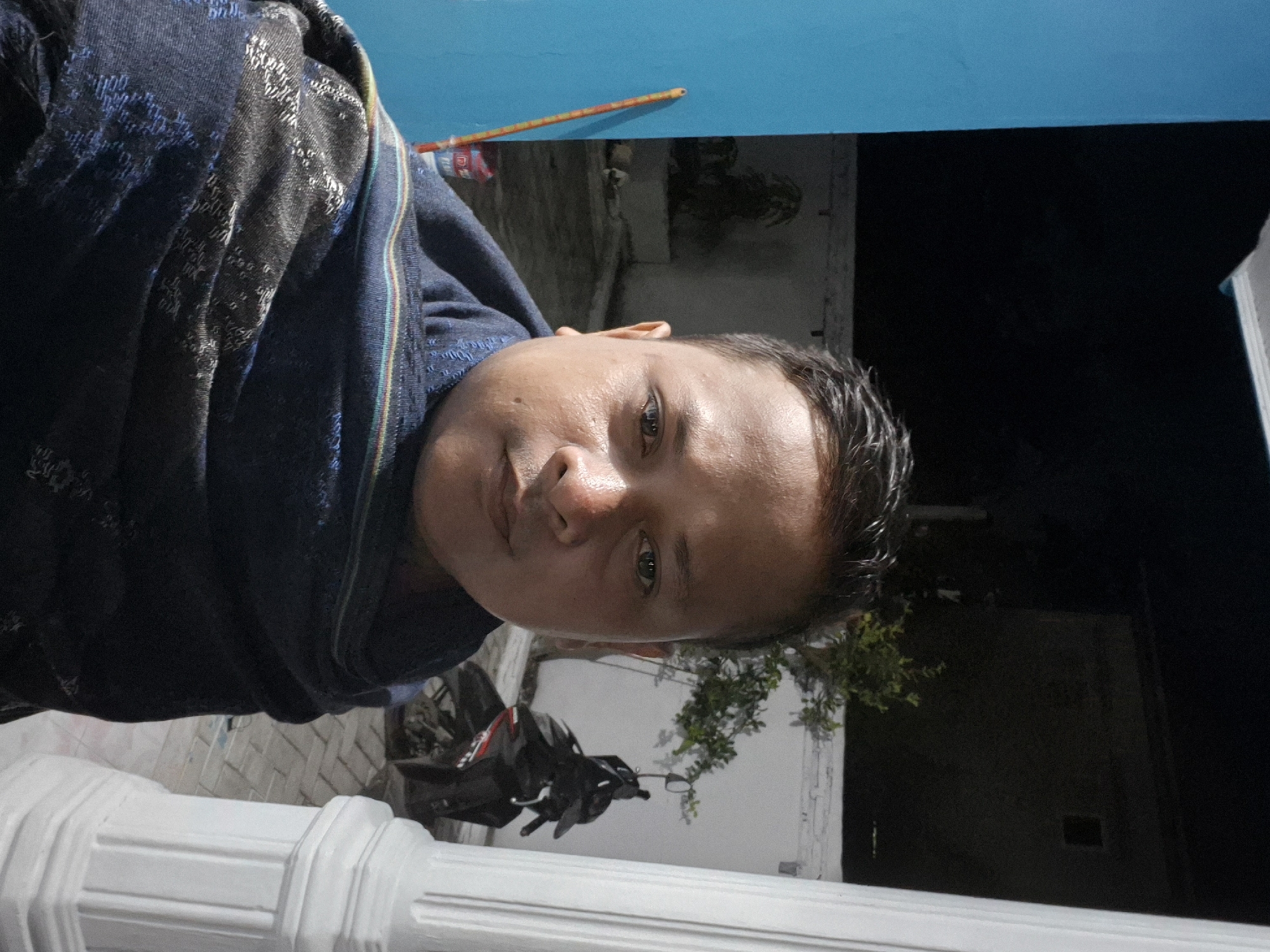 A head-and-shoulders portrait shows a person with tan skin, short dark hair, and a slight smirk. The person is wrapped in a patterned blue and black shawl or garment. The background includes a white, ornate column or railing in the foreground. Behind that is a motorcycle, some foliage, and parts of buildings against a dark sky. A blue surface is partially visible above. The photo was taken outdoors at night, with some ambient lighting. The location cannot be definitively determined to be Lhokseumawe, Indonesia based solely on the visual information provided.