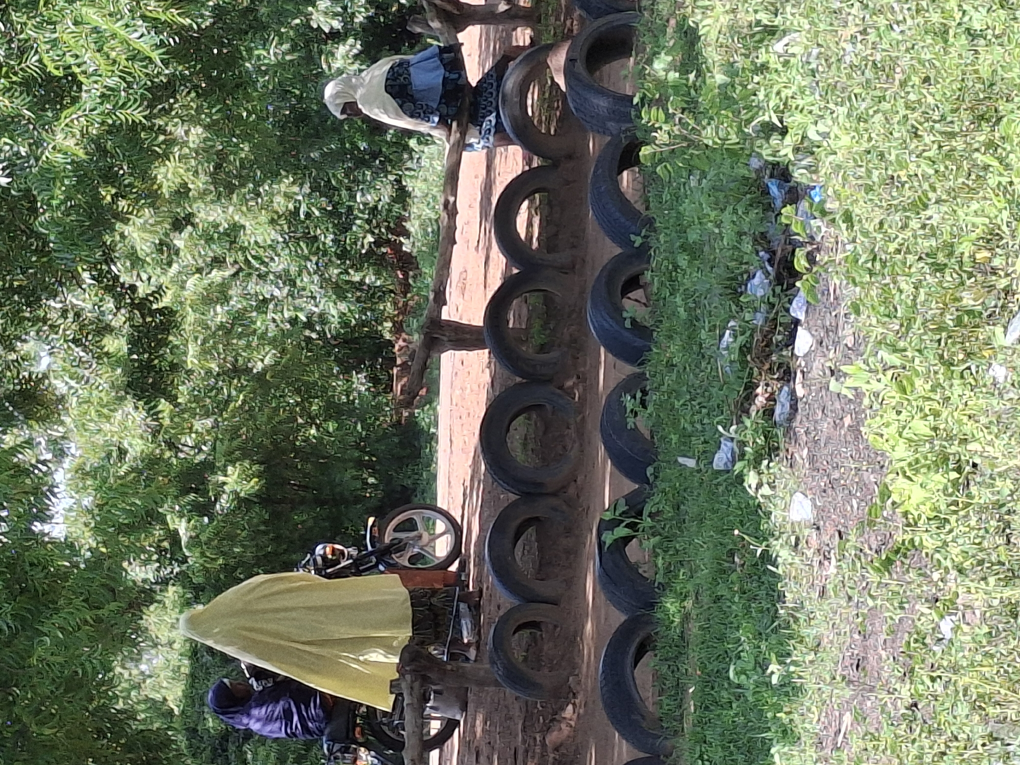 In Dangar, Nigeria, two women are present in an outdoor environment. One woman, wearing a dark hijab, is seated on a motorcycle. Next to her, a second woman is draped in a bright yellow garment. In the background, a motorcycle with a white rim is visible. Further to the right, another woman is seated on a wooden structure, wearing a white hijab and a patterned blue and white dress. The foreground of the image is lined with stacked tires, with grass and some scattered litter in front of them. The scene is surrounded by lush green trees.