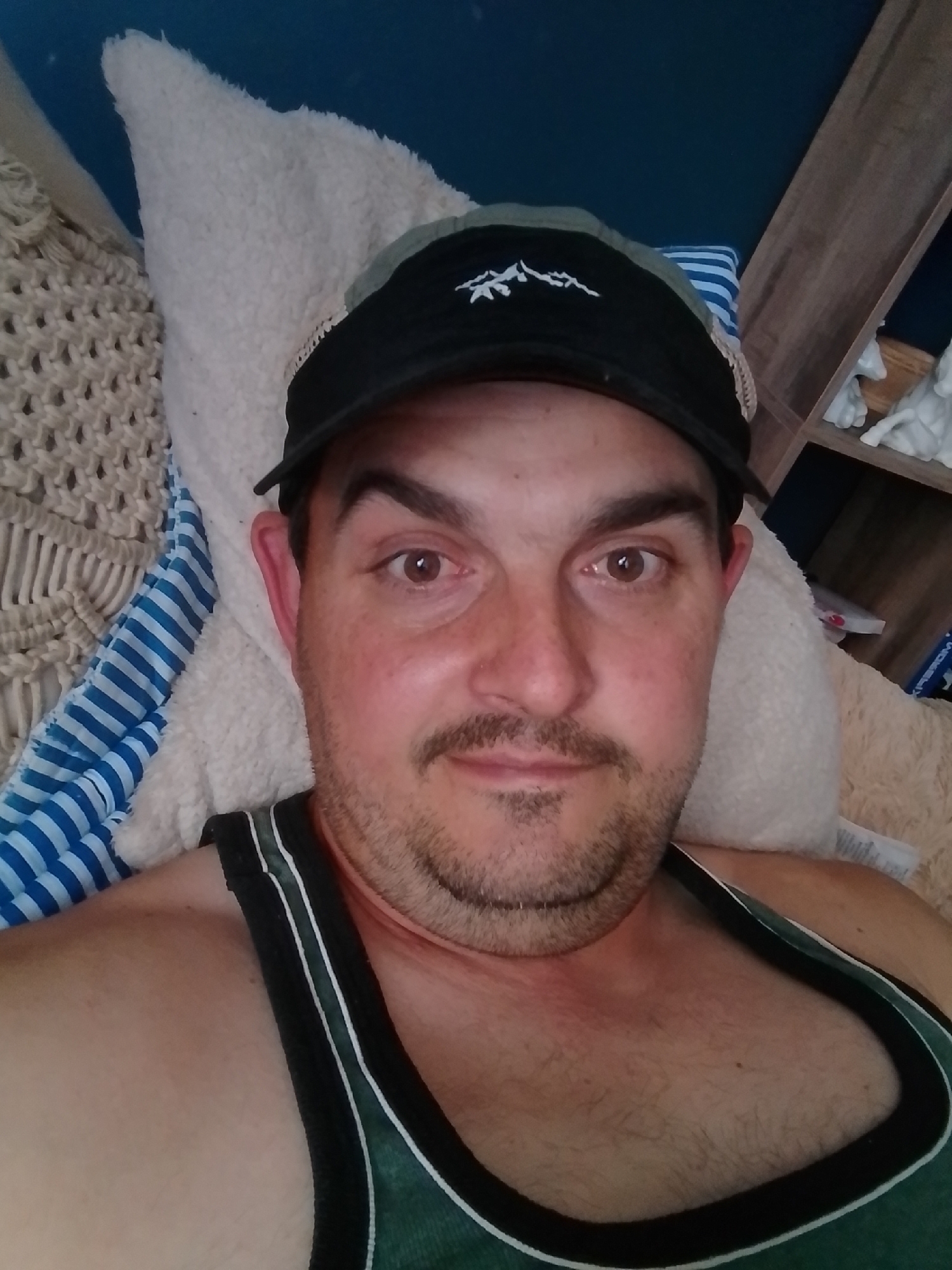 A man is lying down, facing the camera and wearing a black baseball cap and a green tank top. He has light brown eyes, a mustache, and stubble. The man appears to be indoors, lying on a furry white surface with a blue and white striped pillow behind his head. To the right of the man, a wooden shelf unit is visible, with some white decorative items on it. The background is a blue wall. The image does not contain any information that would indicate its location in the city of Wedel, Germany.