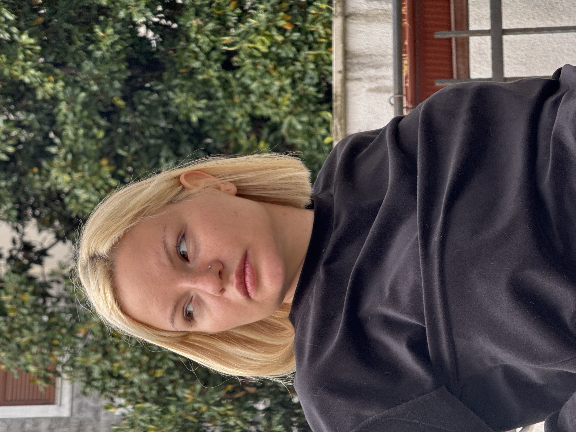 A medium, vertical shot depicts a young adult woman with shoulder-length, blonde hair. She wears a black hooded sweatshirt and has a nose piercing. The woman is looking downward and to the left. The background includes green foliage and a building with a window. The location is unspecified, and there is no information regarding a specific location within Budva, Montenegro.