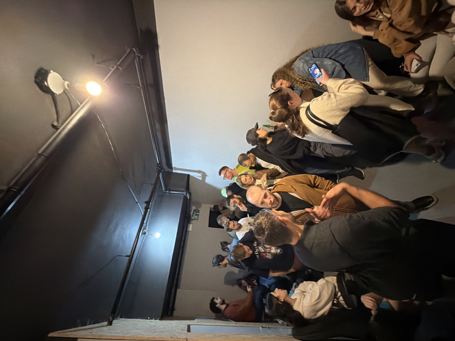 The image displays an indoor social gathering within a dimly lit venue in Tel-Aviv, Israel, as indicated by the Hebrew "יציאה" (Exit) sign visible above a doorway in the background-left. The space features a black ceiling with exposed metal conduits and two track lighting fixtures, along with a single bare incandescent bulb providing illumination in the back-left. The right wall is plain light grey, while the left wall has a textured, possibly concrete or wood-paneled surface.
Approximately 25-30 individuals, predominantly young adults, are present. Most are dressed in casual attire, including sweaters, hoodies, t-shirts, and caps. People are observed standing, sitting, and interacting in various groups. In the foreground, two men face each other, engaged in conversation; the man closer to the viewer, wearing a dark top, gestures with his right hand towards the other man, who wears a brown hoodie. To their right, a woman in an off-white sweater holds a smartphone, viewing its screen, with a dark tote bag slung over her shoulder. Adjacent to her, a woman with curly hair, wearing a denim jacket, is seated or leaning against a low surface, also looking at a smartphone.
In the midground, individuals are mingling, with several standing close to the right wall. One woman wears a black cap and holds a green bottle, possibly a beverage. In the background-left, a man in a red-brown sweater is seated on a ledge, conversing with others seated below him. Various other individuals are distributed throughout the scene, some observing, some in conversation, and some appearing to use their mobile devices.