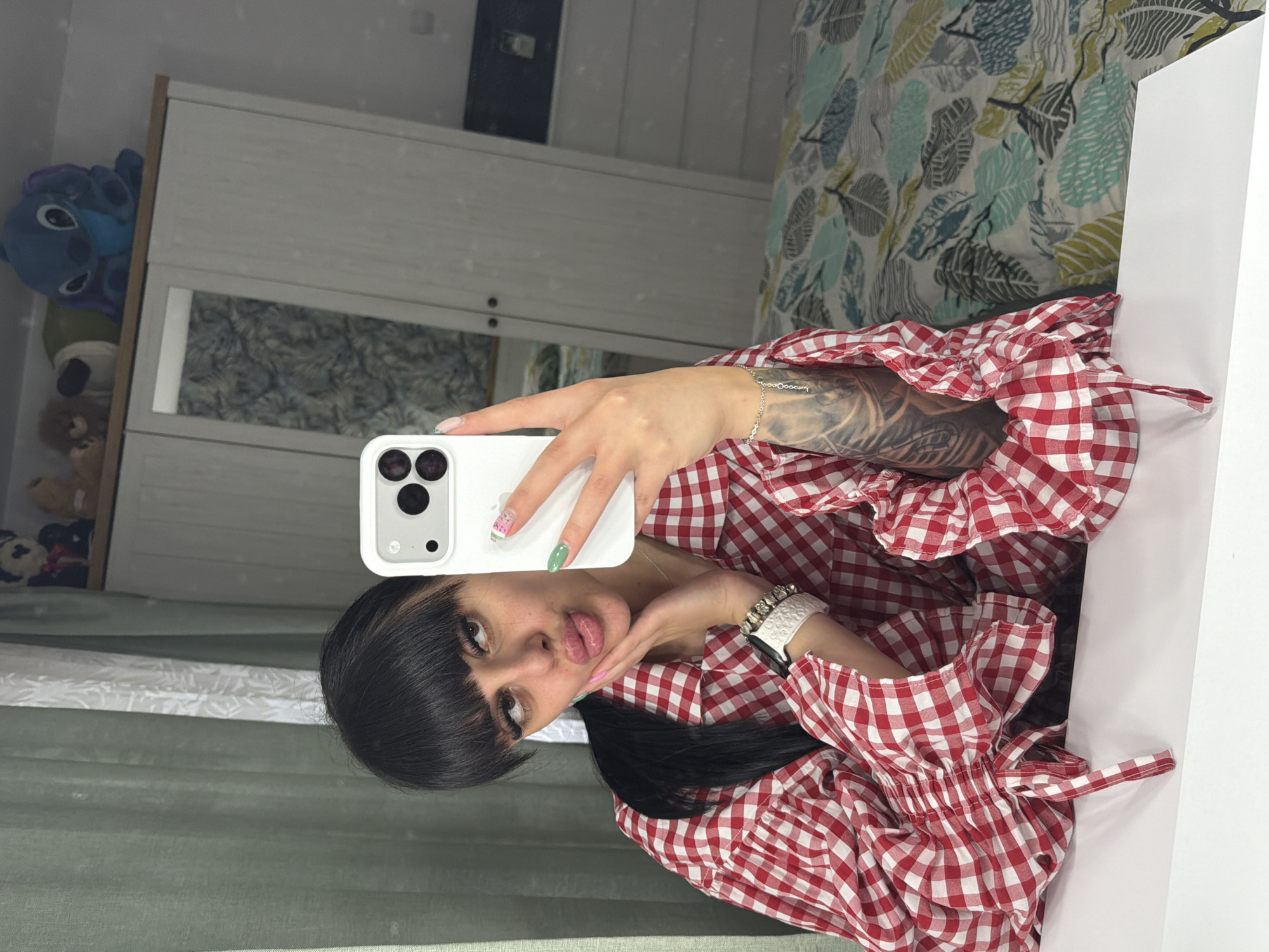 A young woman captures a mirror selfie in what appears to be an indoor setting in Plovdiv, Bulgaria. The image is oriented sideways, with the subject leaning to her left.
The woman has dark hair with bangs and is wearing a red and white checkered top with ruffled details. She is making a playful facial expression, sticking her tongue out slightly. She holds a white smartphone with multiple camera lenses in her left hand, which is adorned with manicured nails featuring various colors and designs, including pink with patterns and a solid green nail.
Her left arm, partially visible, displays a prominent tattoo that includes text, appearing to spell "Doocany" or similar, and she wears a delicate silver chain bracelet. On her right wrist, she has a white smartwatch.
The reflections in the mirror reveal details of the room. To her left, a white cabinet or dresser is visible, with several stuffed animals, including a blue one resembling Stitch, placed on top. To her right, a bed is visible with a duvet or comforter featuring a distinctive pattern of teal, green, and grey leaves. A green curtain or textile is visible in the lower part of the reflected scene. The overall impression is a casual, personal moment captured in a home environment.