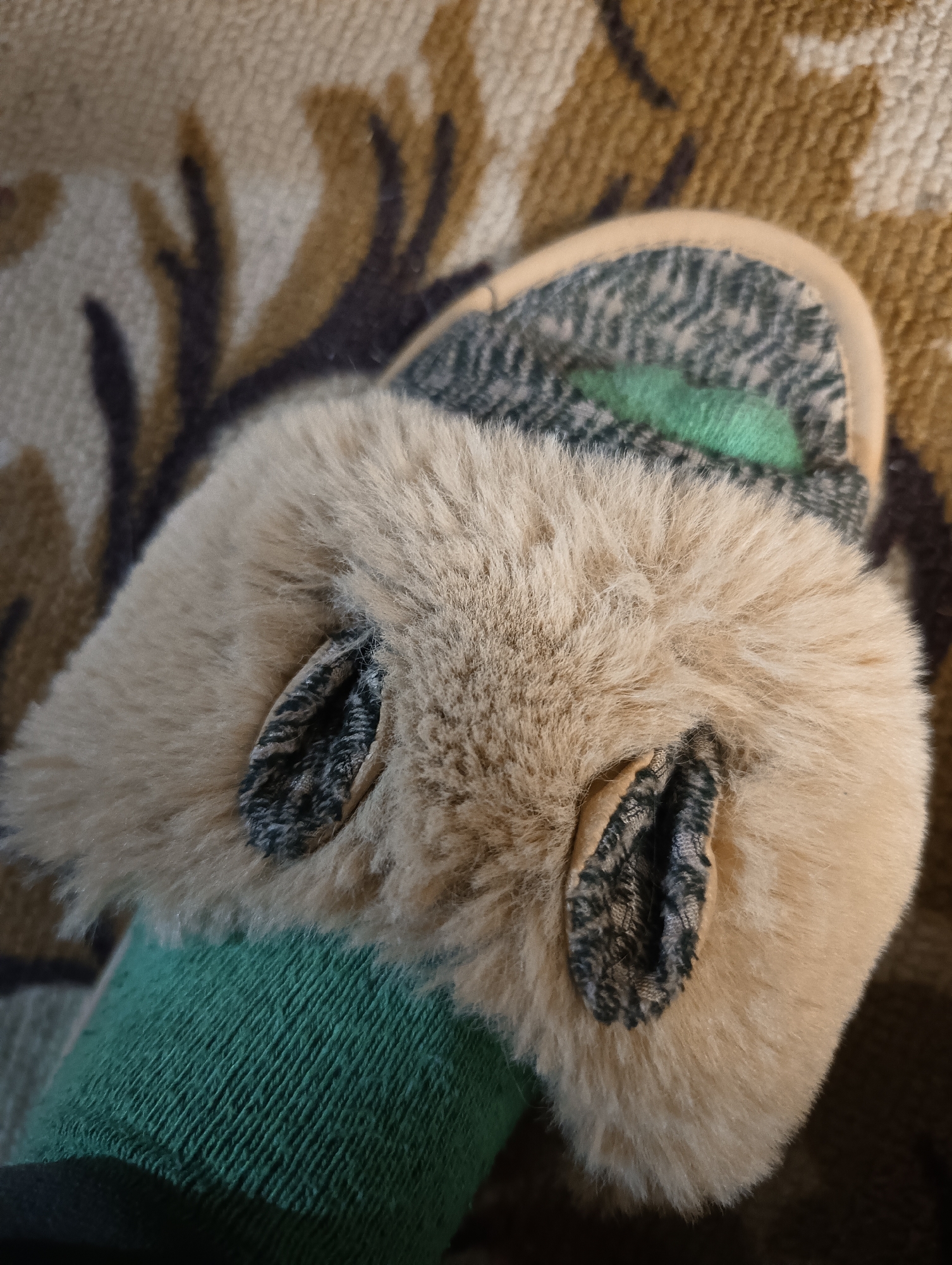 The close-up image features a person's foot, encased in a vibrant green sock, resting within a fluffy slipper. The slipper is primarily light brown or tan, covered in soft faux fur that gives it a plush appearance. Two small, ear-like elements, made from a patterned fabric of grey and black hues, protrude from the top of the slipper. A thin, light beige trim frames the opening of the slipper.
A glimpse of a darker green lining or material is visible deeper within the slipper, near the toe area. The background consists of a blurred fabric, possibly a blanket or carpet, displaying an abstract, organic pattern in shades of brown, beige, and dark grey, reminiscent of branches or foliage. The overall impression is one of comfort and relaxation in an indoor setting. No specific activity is depicted other than the foot at rest. There are no discernible indications of the time of day, weather, or visible text. The location context for the image is Biliaivka, Ukraine.