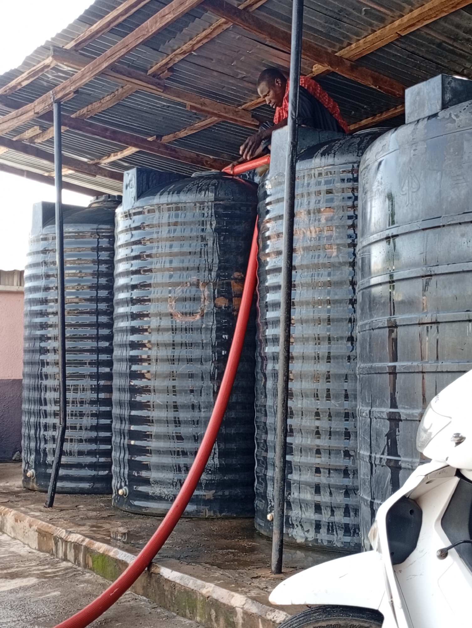 A person wearing a red scarf around their neck and a black shirt is seen working on top of a large black water tank. They are reaching down into the tank, possibly to adjust or clean something. In front of the water tanks, a red hose is coiled on the ground and extends upwards towards the tanks. The water tanks are cylindrical and appear to be made of plastic. They are lined up next to each other under a corrugated metal roof supported by wooden beams and a metal pole. The ground around the tanks is wet and appears to be concrete. A portion of a white motorcycle is visible in the lower right corner of the image. The scene suggests an outdoor utility area, likely for water storage.