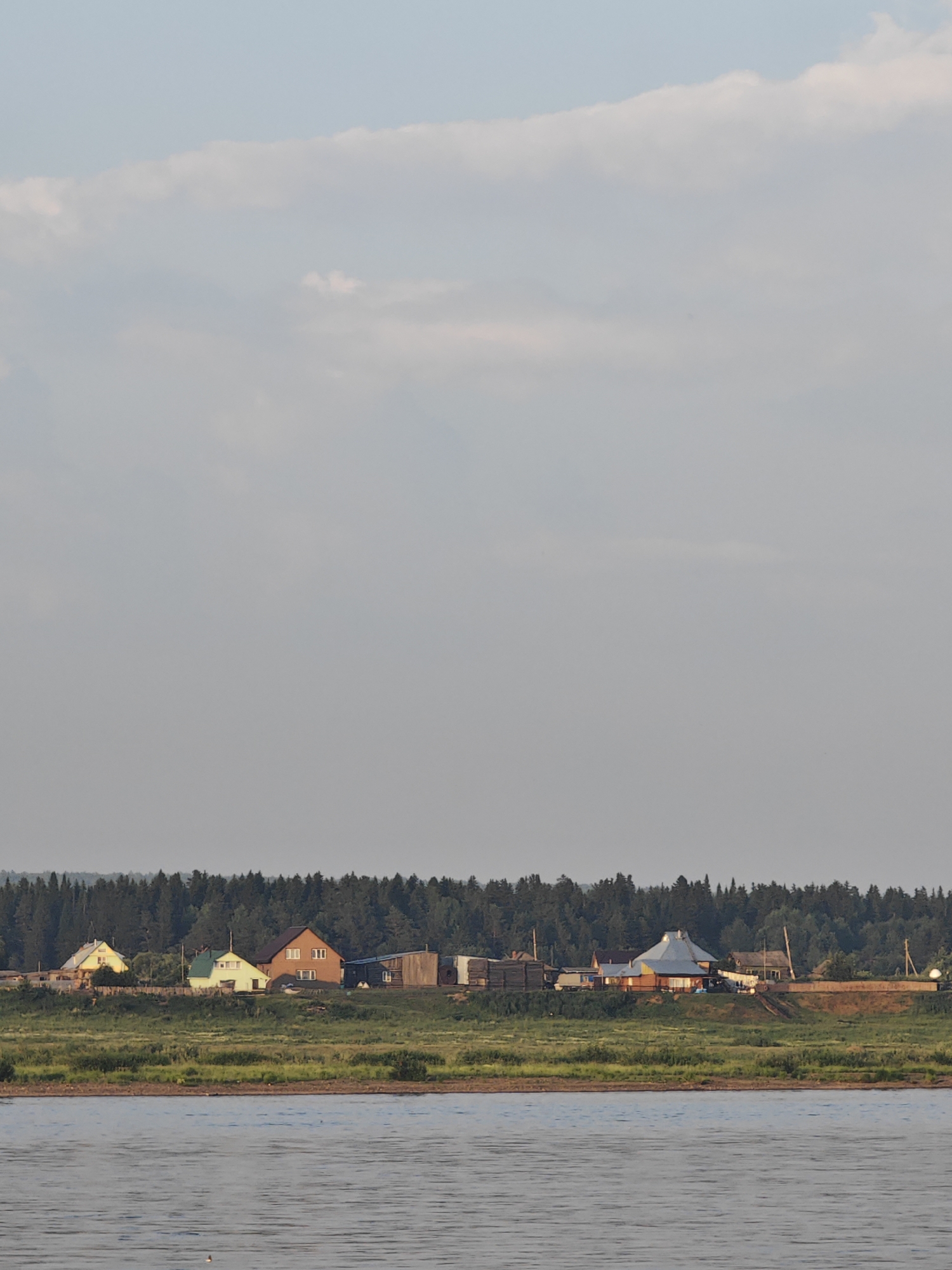 The image depicts a rural scene with a wide river in the foreground, reflecting the soft light of the sky. In the middle ground, a densely forested area forms a dark green horizon line. Along the crest of the bank, overlooking the river and forest, is a small settlement of houses. These structures appear to be typical rural dwellings, some with visible chimneys and varied rooflines. The architecture suggests a temperate climate. The overall atmosphere is serene and suggests a peaceful, remote location. The location is identified as the city of Lesosibirsk, Russia, a city known for its timber industry and location in Siberia, which aligns with the natural landscape depicted.