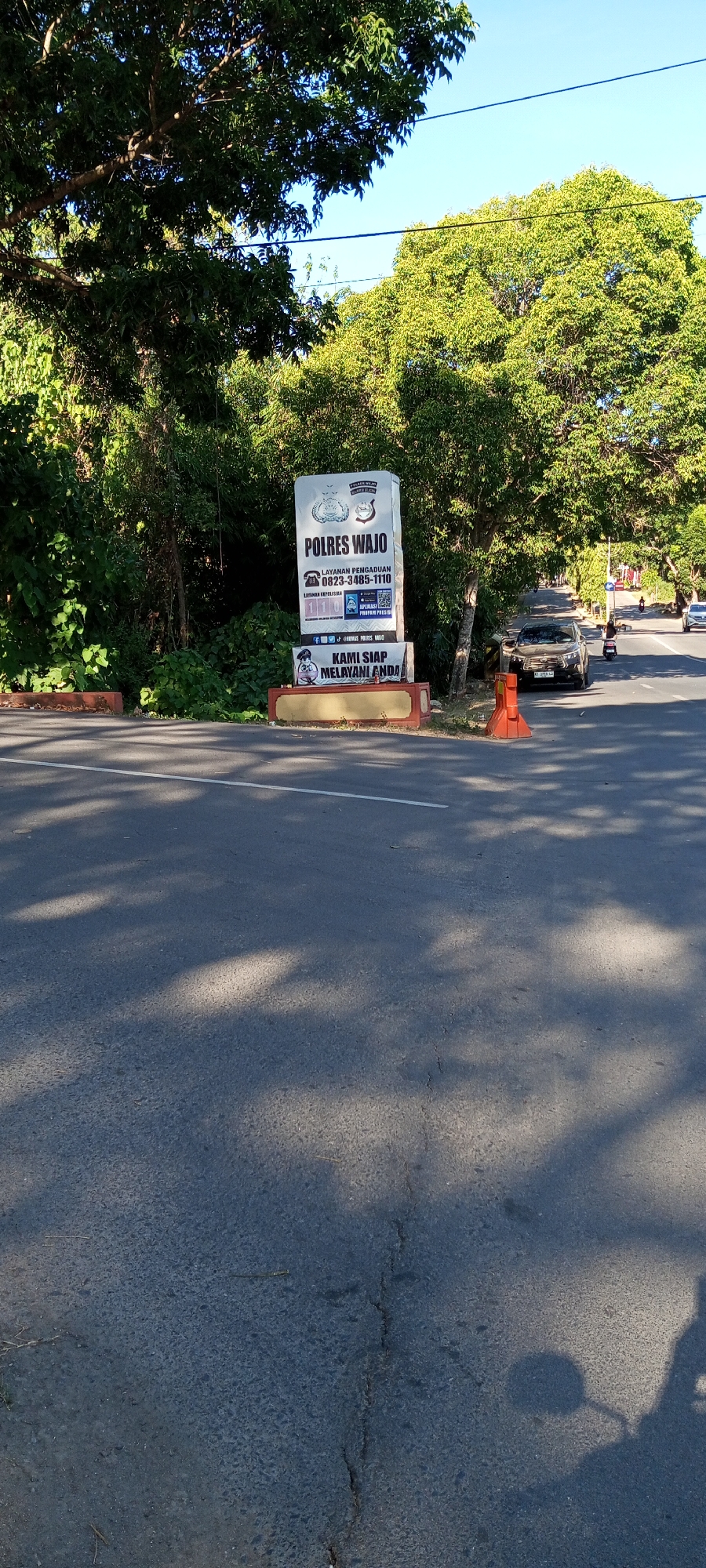 The image shows a street scene in Bulupabbulu, Indonesia. A tall, white sign with text and graphics is prominently displayed on the right side of the road, next to a wooded area. The sign reads "POLRES WAJO" (Wajo Police) and provides contact information including a phone number and an app name. Below the main sign, a smaller sign features a cartoon police officer and the text "KAMI SIAP MELAYANI ANDA" (We are ready to serve you). Orange traffic cones are placed on the side of the road. In the background, trees line the street, and traffic, including cars and motorcycles, can be seen on the road. The sky is clear and blue. The road surface is asphalt, with shadows of trees cast across it.