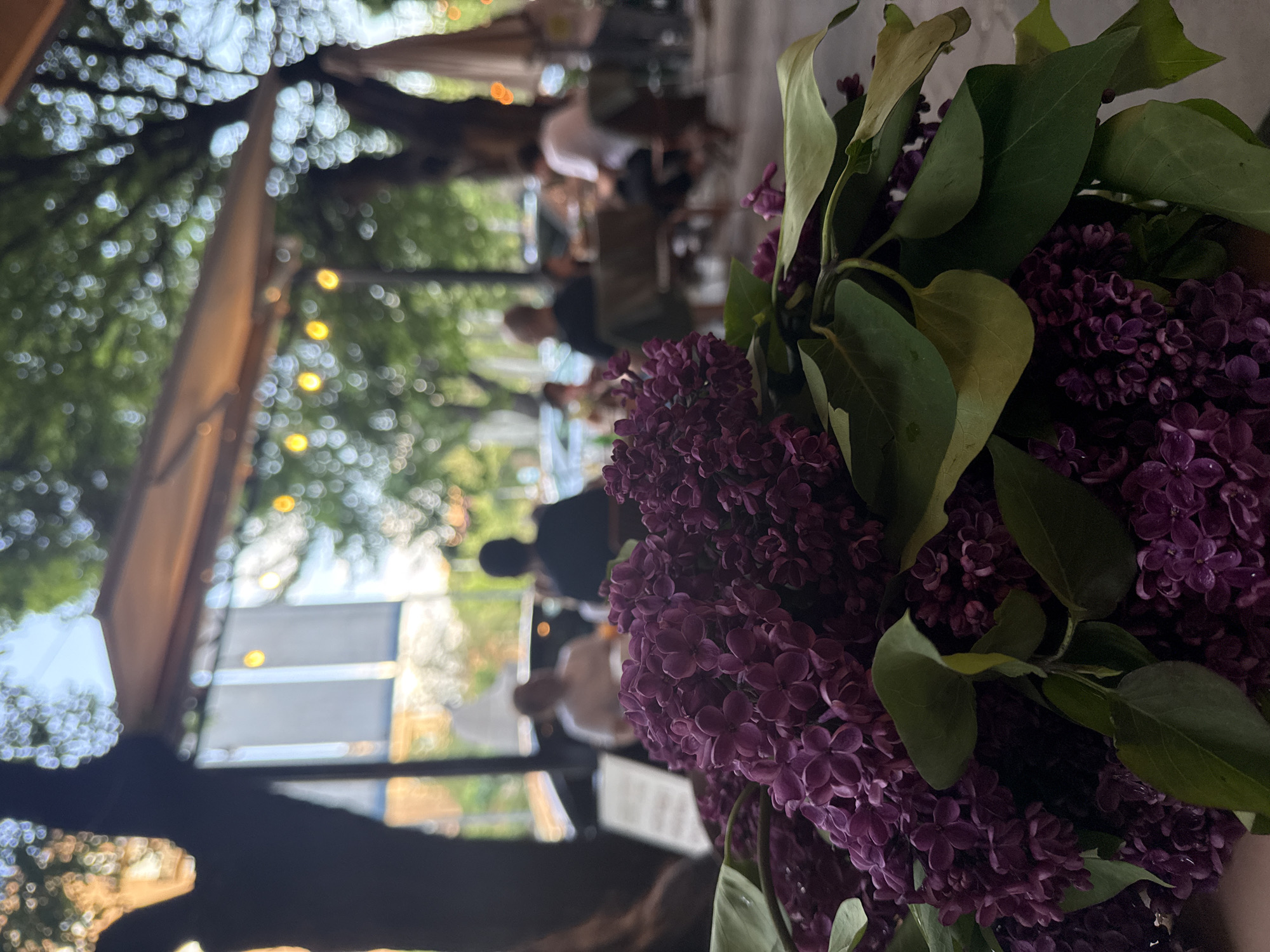 A close-up, eye-level shot shows a bouquet of purple lilacs in the foreground, partially obscuring the background. The background is a blurred outdoor restaurant scene, presumably in the city of Yerevan, Armenia. People are seated at tables, engaging in activities such as eating and conversation. String lights are visible overhead, suggesting a relaxed and ambient atmosphere. The image is slightly tilted, giving it a dynamic feel. A menu or document is partially visible below the flowers.