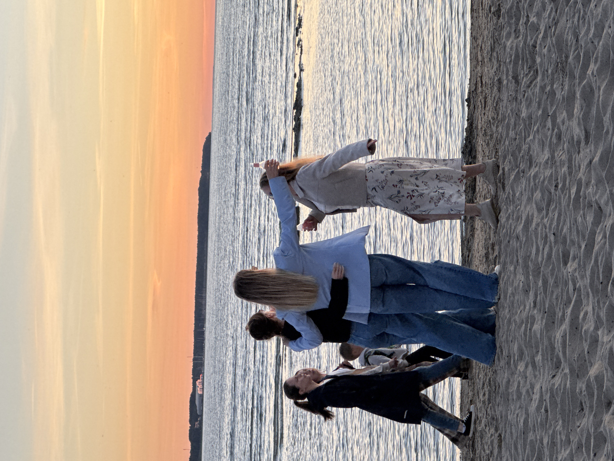 A group of four people, two women in the foreground and two in the background, are standing on a sandy beach by the sea at sunset. The two women in the foreground are facing away from the camera, with one in a light blue blazer and the other in a cream-colored jacket and a floral print skirt. They are holding bottles. The woman in the blue blazer has her arm around the other woman's shoulder. In the background, two other individuals, a woman and a child, are walking away from the camera. The sky is a gradient of orange and yellow, indicating sunset. The sea is calm with gentle ripples. In the distance, a tree line with some buildings and a marina can be seen along the horizon. The image appears to have been taken in Tallinn, Estonia, likely along the coast such as Pirita or Kakumäe.