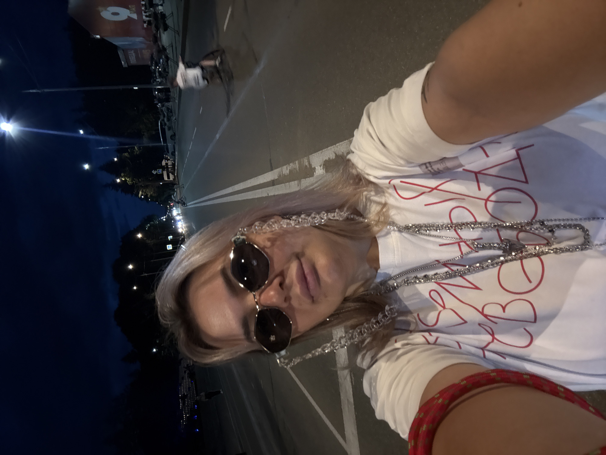 In this selfie, a person with blonde hair and sunglasses is depicted outdoors at dusk. They are wearing a white t-shirt with red lettering and several necklaces, including one with clear beads and another with silver chains. A red and gold bracelet is visible on their arm. The background shows a dark sky with scattered lights, suggesting an urban or suburban setting at night. The ground appears to be asphalt with white lane markings, indicating a road or parking lot. A street light is prominently visible in the upper left corner. The overall mood is casual and appears to be a self-portrait taken during an outing. The red text on the t-shirt is partially visible and appears to form stylized letters, but it is not fully legible.