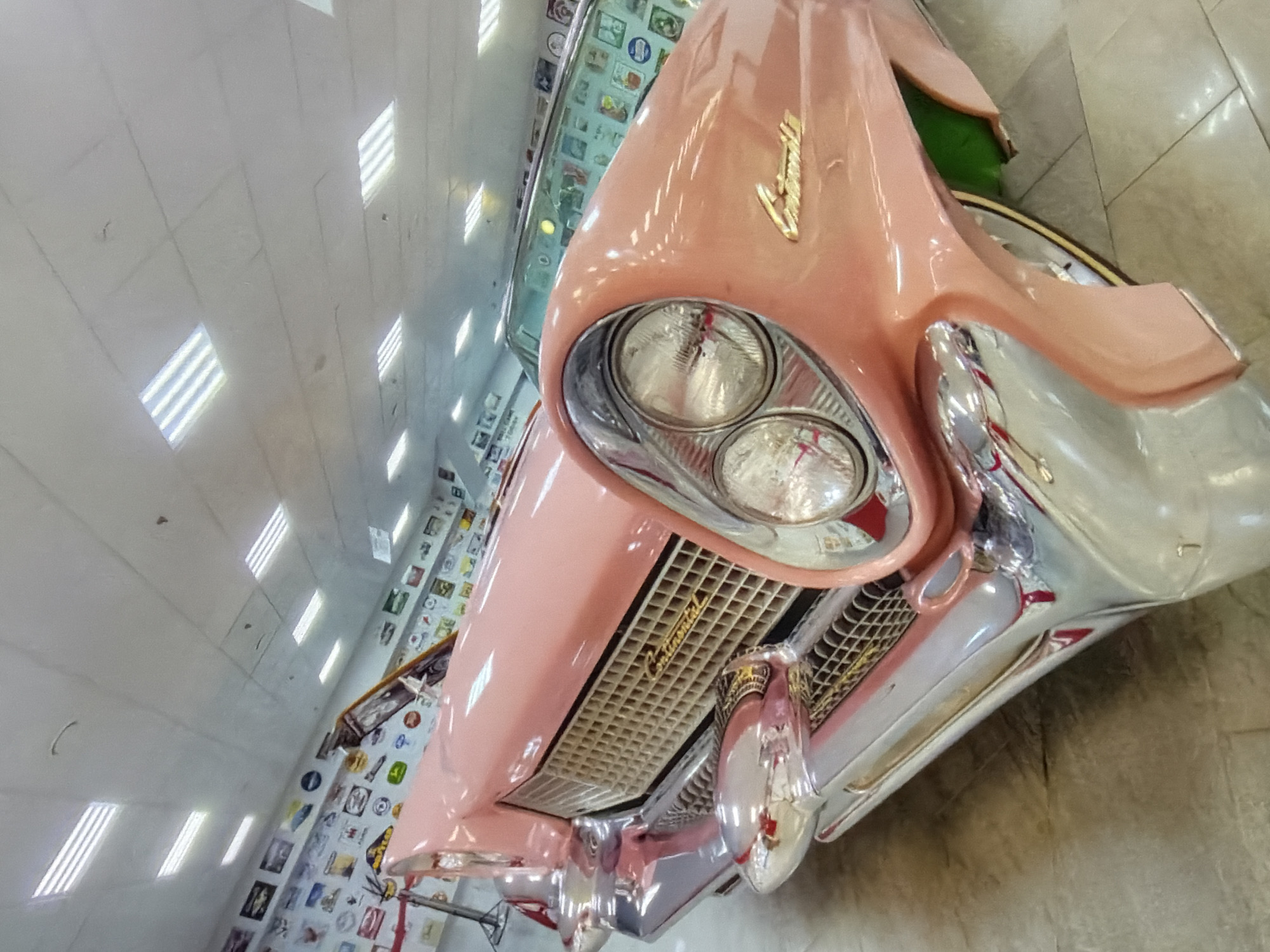 A low-angle, close-up shot depicts a pink vintage car, likely a Cadillac, inside an indoor space in the city of Unknown, Unknown. The car's front end, particularly the headlights, grill, and chrome bumper, dominates the view. The setting appears to be an automotive museum, with a wall decorated with photographs or memorabilia visible behind the car. The ceiling is tiled with recessed lighting fixtures. The floor has a tiled surface with a tan coloring. The overall lighting in the scene is bright, reflecting off the car's polished surfaces.