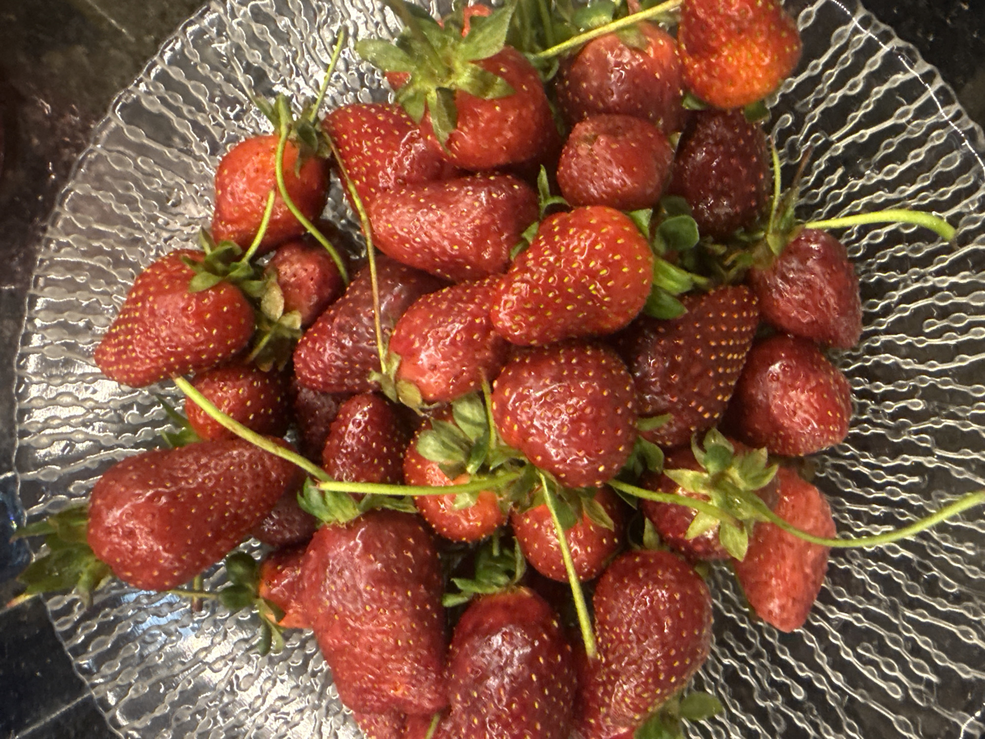 A close-up shot presents a glass bowl filled with fresh strawberries. The strawberries are a deep red color and appear ripe, with green stems attached to some. The bowl has a textured pattern of concentric lines. The overall setting appears to be indoors. The location is unspecified.