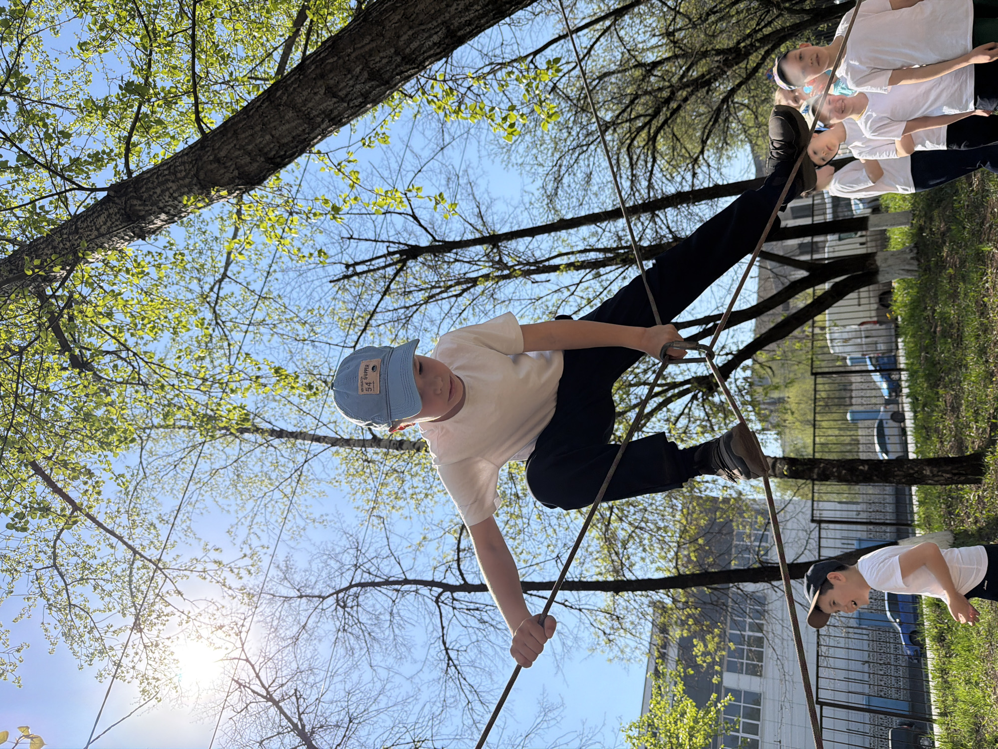 A young boy is navigating an obstacle course made of ropes and trees at a park in Izhevsk, Russia. The course is set against the backdrop of a sunny day, with trees in the background and a building visible in the distance. The boy is wearing a white t-shirt, dark pants, and a blue cap, and he is using his hands and feet to balance on the ropes. Other children and adults are present in the surrounding area.