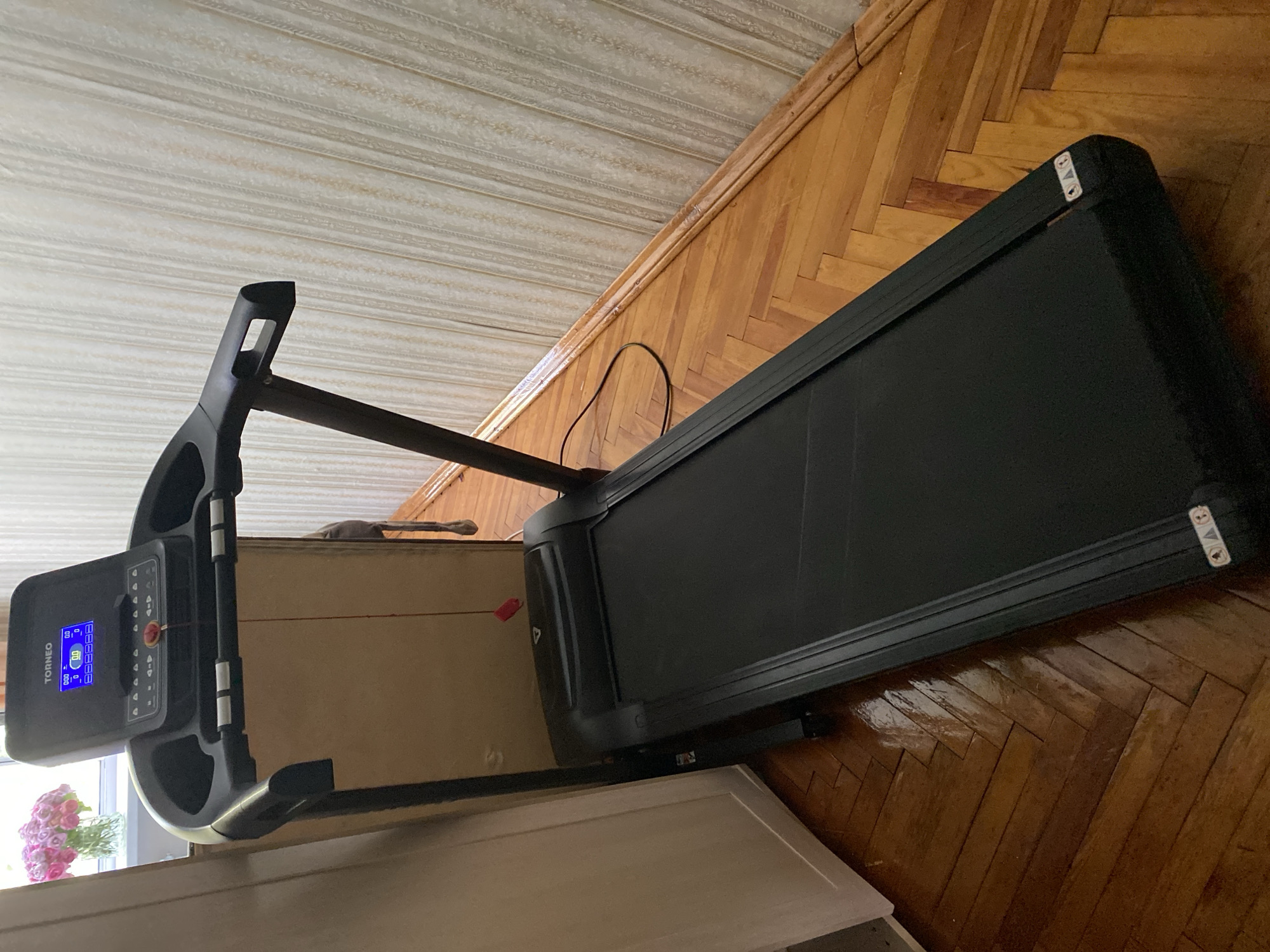 The image shows a black treadmill with a digital display on a wooden floor. The treadmill is positioned in what appears to be a room with patterned wallpaper. To the left of the treadmill, a portion of a white door or cabinet is visible, and a vase of pink flowers sits on a windowsill in the background. A red safety clip is attached to the console of the treadmill. The floor is made of polished hardwood arranged in a herringbone pattern. The overall scene suggests an indoor setting, likely a home or gym. There is no indication of the specific location in the city of Lomonosov, Russia, beyond it being an indoor environment.