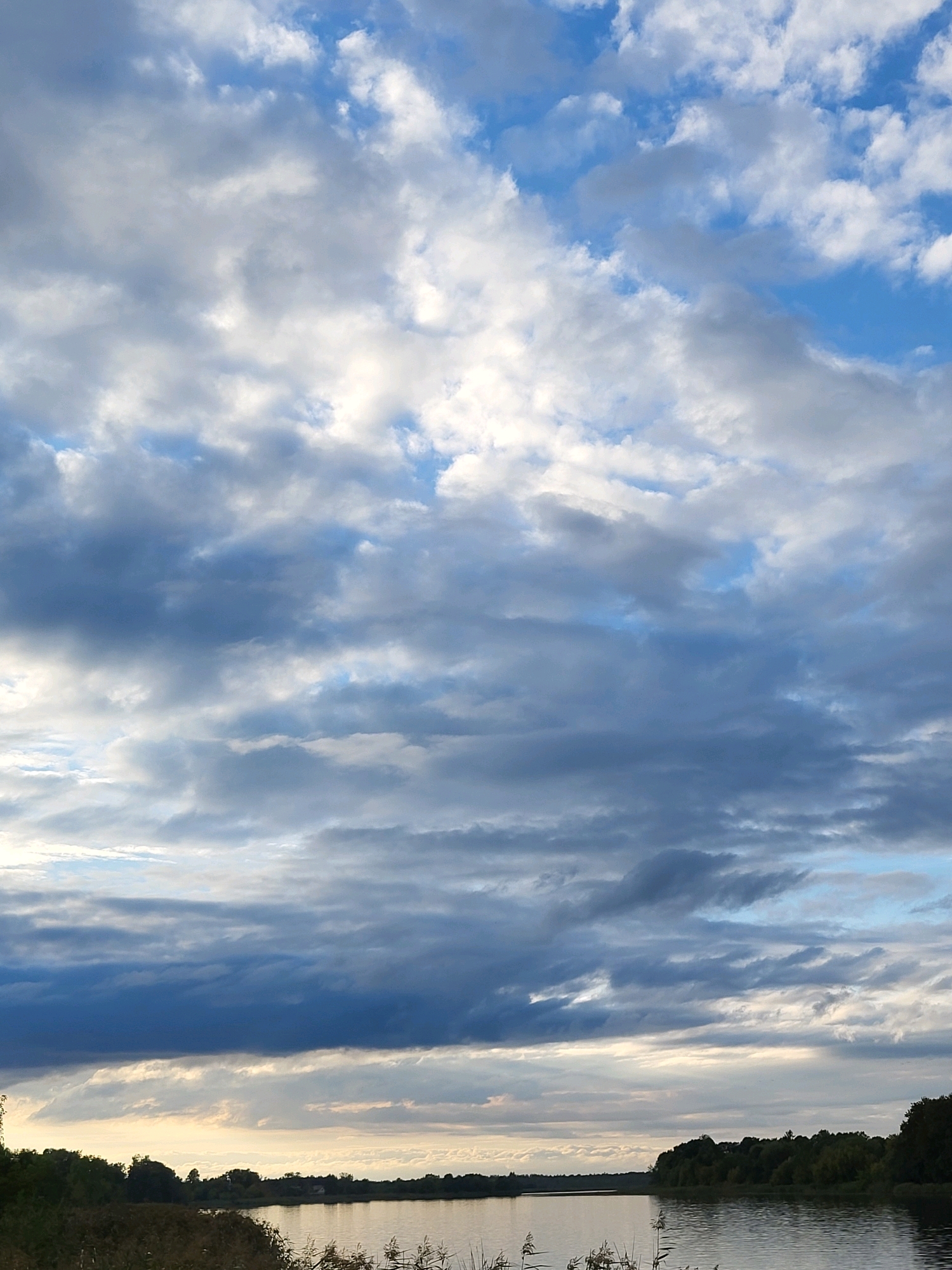 The image shows a body of water, likely a river, on a cloudy day. The water is calm and reflects the sky. Along the far bank of the river, there is a line of trees, and in the distance, a few houses are visible. The sky is filled with various cloud formations, ranging from light and wispy to darker, more ominous storm clouds. The overall impression is one of a serene, natural landscape under a dramatic sky. The location is identified as Kalnciems, Latvia.