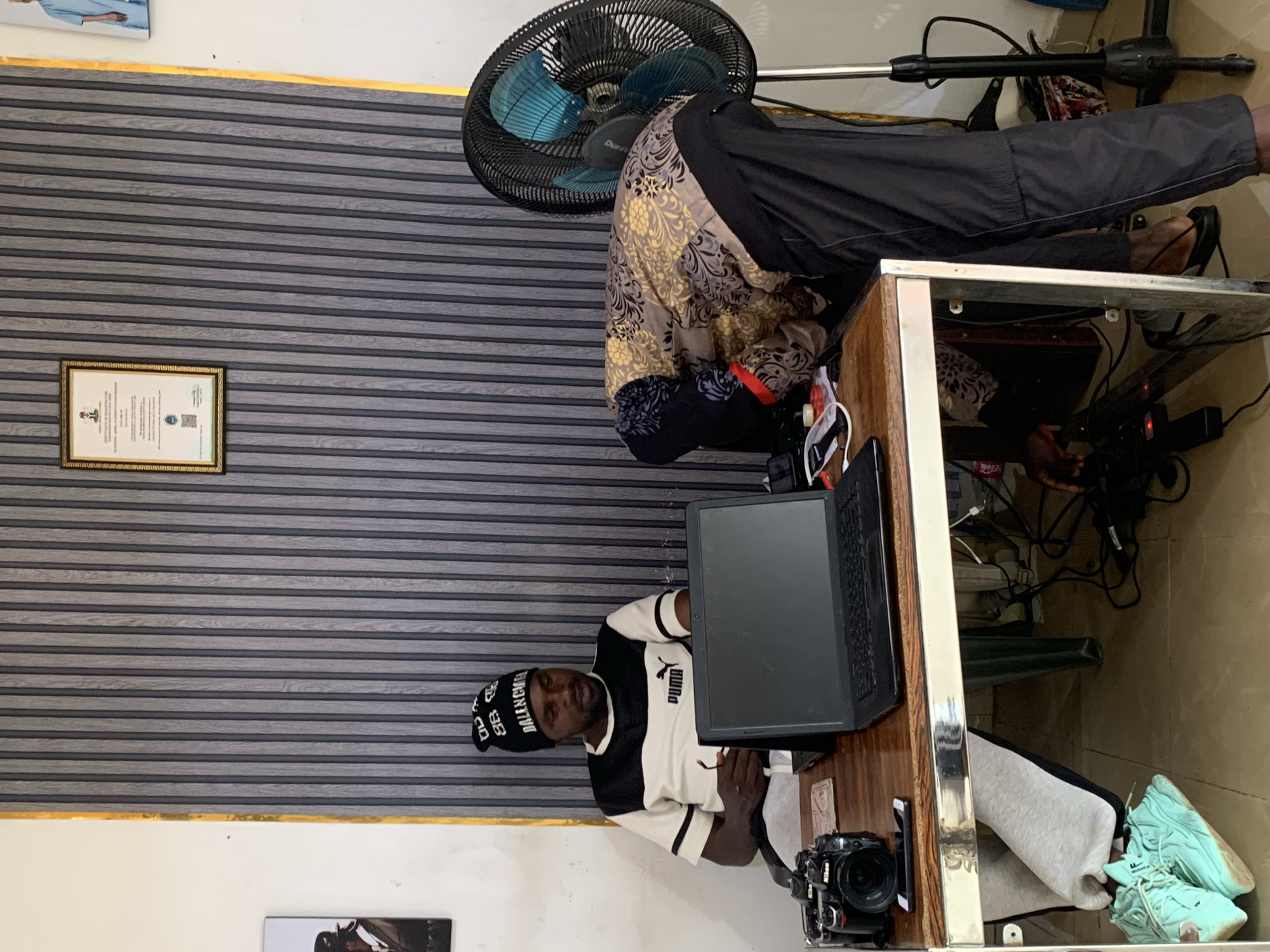 The image depicts an indoor scene, possibly in Jos, Nigeria, given the context provided. A man wearing a black beanie with white lettering and a black and white Puma t-shirt is seated at a wooden desk, looking down. He is holding a pair of glasses in his right hand. On the desk in front of him is a black laptop, a Nikon camera, and a smartphone. To the right of the desk, another person, whose face is not visible, is bent over, wearing a patterned jacket and black pants. A black oscillating fan is positioned behind this person. The wall behind them features a striped textured pattern and a framed certificate hangs on the wall. Underneath the desk, a pair of light blue sneakers are visible. Various cables and electronic devices are scattered on the floor beneath the desk. The lighting suggests an indoor setting, likely during daytime.