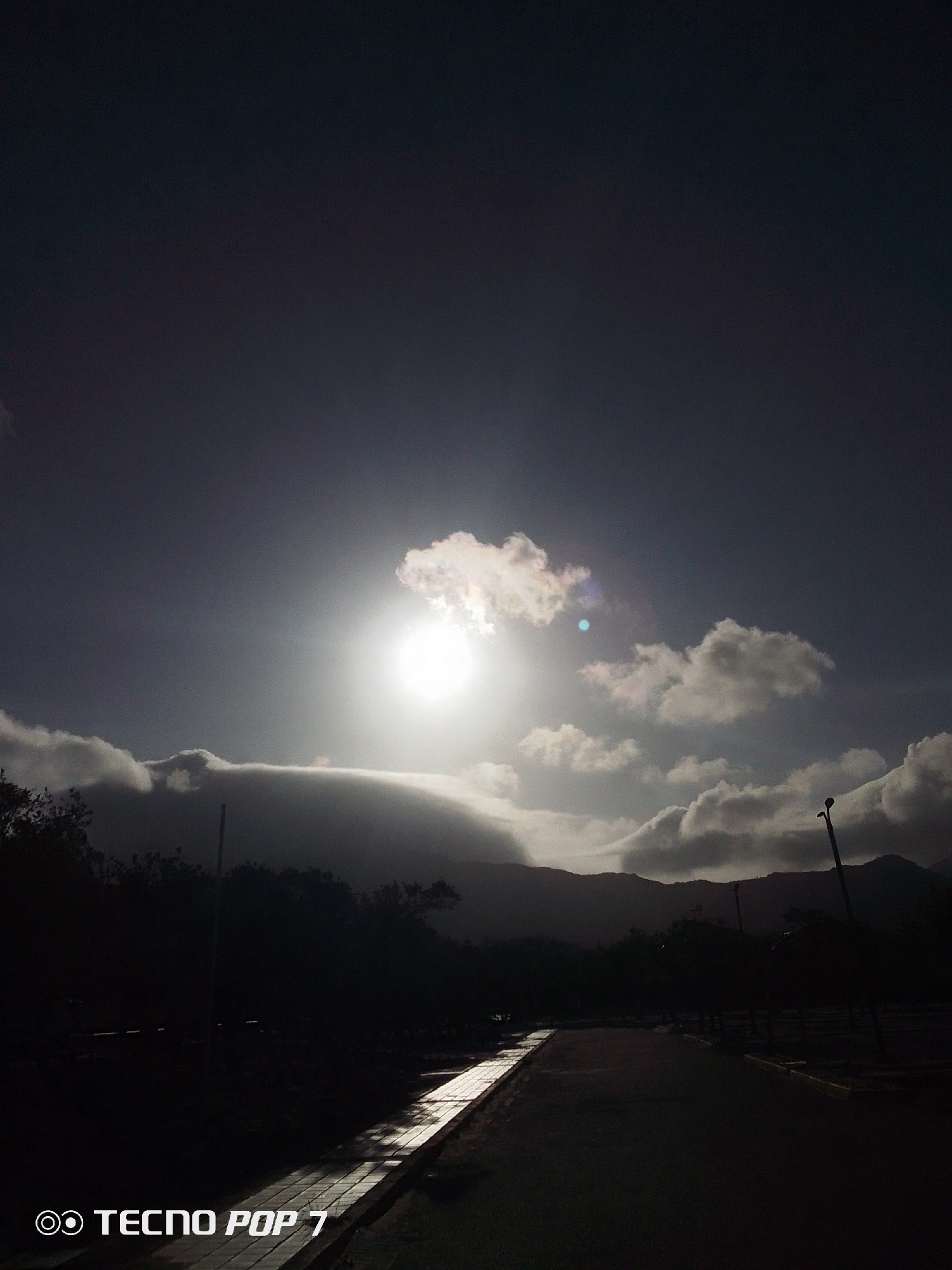 The image depicts a low-angle view of a road in Pedro González, Venezuela, under a bright sky. The sun is the focal point, surrounded by clouds, creating a high-contrast scene where the lower portion is obscured in shadow. In the foreground, the road surface appears dark, though a segment reflects light, revealing a paved or tiled edge. Beyond the road, silhouettes of trees and mountainous terrain are visible. A few utility poles stand on the right, while the name "TECNO POP 7" is displayed in the lower left corner.