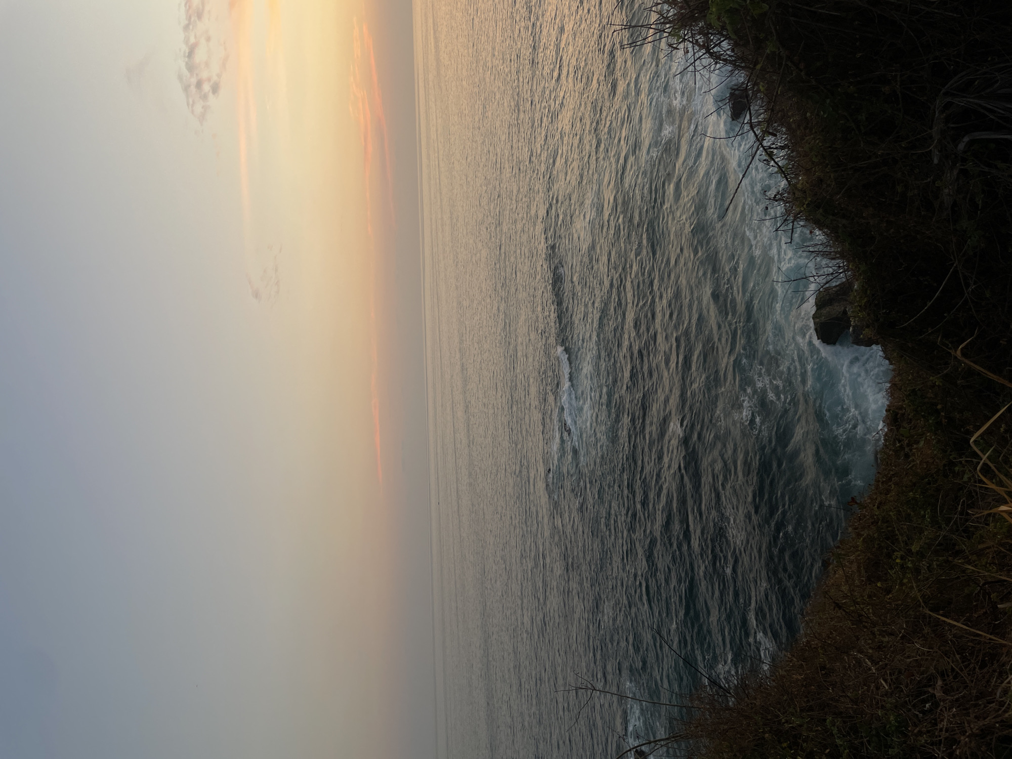 The image captures a view from a cliff overlooking the ocean, likely at sunset or sunrise due to the soft orange and grey hues in the sky. The sea is visible and waves are breaking against the rocks at the base of the cliff. The cliffside is covered in dense vegetation. The location is likely in Puerto Escondido, Mexico. There are no people visible.