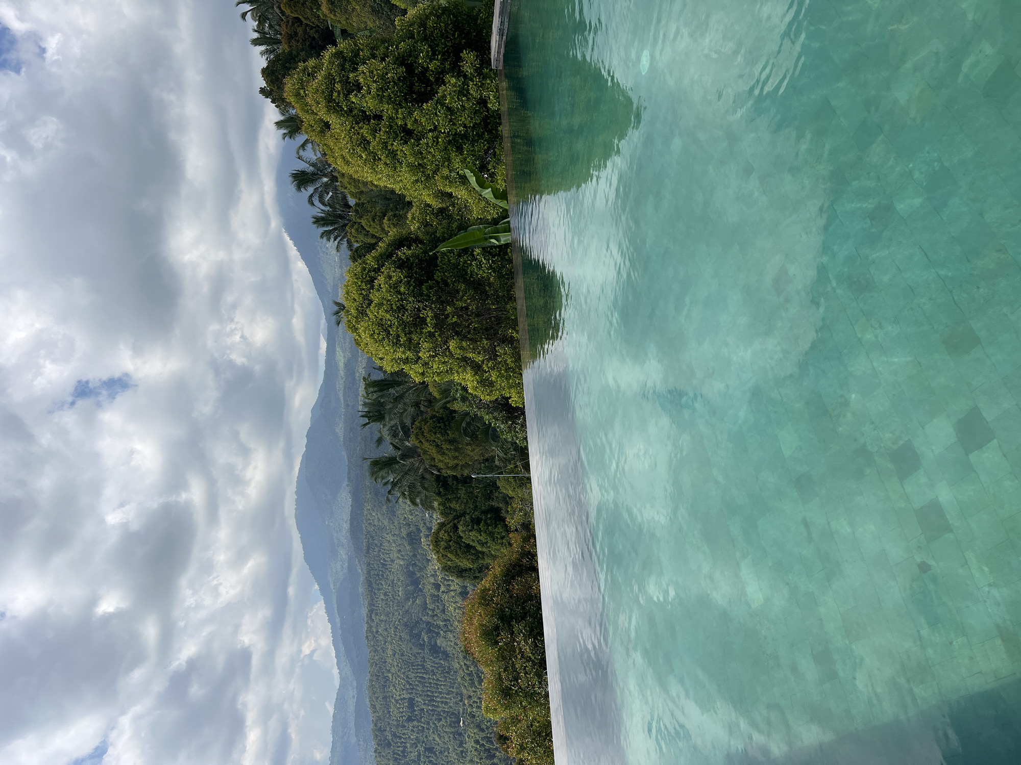This is a scenic photograph of an infinity pool overlooking a lush, mountainous landscape in Busungbiu, Indonesia. The pool's turquoise water fills the foreground, its surface reflecting the sky and surrounding greenery. Beyond the pool, rolling hills covered in dense vegetation stretch towards distant mountains. The sky is partly cloudy, with dramatic grey clouds interspersed with patches of blue. The overall impression is one of serene natural beauty and tranquility. There are no people or animals visible in the image.