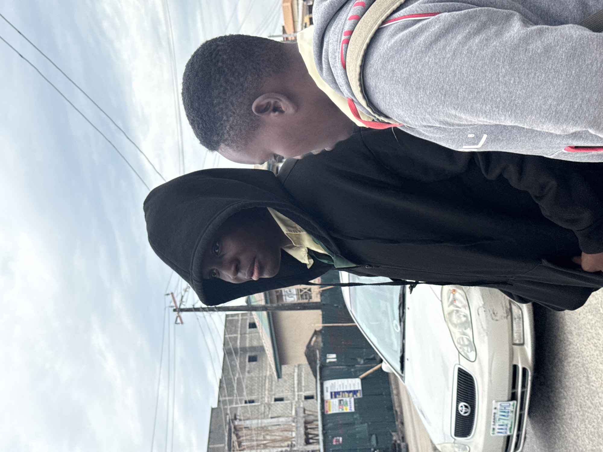 Two young men stand on a street beside a parked white car. One man, with dark skin and short hair, wears a grey hoodie with a beige strap and red accents. He leans towards the other man. The second man wears a black hooded jacket, obscuring his face, but his dark eyes and a hint of a yellow shirt are visible. Behind them, a partially constructed building and an electricity pole are visible. The car's license plate reads "AAA 744 HD." The buildings suggest an urban setting in Ado, Nigeria.