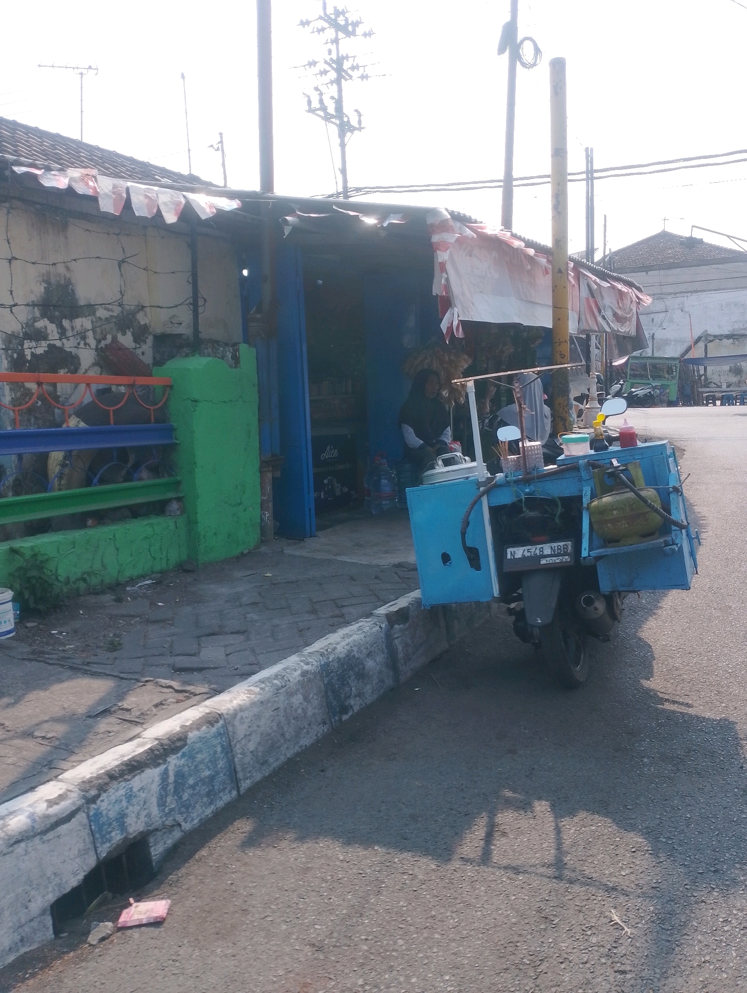 The image depicts a street scene in Probolinggo, Indonesia, likely near a food stall. A modified motorcycle, fitted with a blue cart, is parked on the asphalt road. The cart appears to be a mobile food vendor setup, containing a gas cylinder, cooking utensils, and various containers. A person, possibly the vendor, is seated in the background, partially obscured by the stall's awning. The stall itself is constructed with a combination of brick walls and corrugated metal roofing, covered by a red and white striped tarp. A brightly painted fence, with green, blue, and orange sections, borders the area in front of the stall. The ground is paved with brickwork in some areas and asphalt in others, with a concrete curb and a drainage ditch running along the road. Electrical poles and wires are visible in the background, indicative of an urban or semi-urban environment. The lighting suggests it is daytime, with the sun casting shadows across the scene.