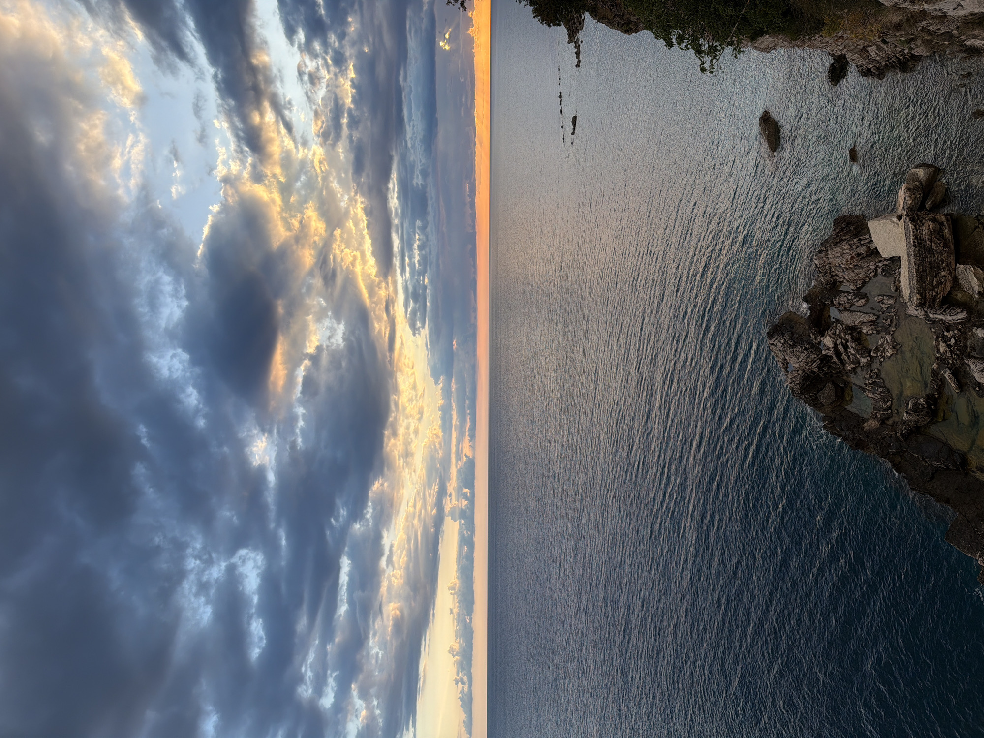 The image depicts a panoramic view of the Adriatic Sea from the coastal cliffs of Petrovac na Moru, Montenegro. The perspective is a high-angle shot, capturing the contrast between the dynamic sky and the tranquil sea.
The sky, occupying the left portion of the image, is filled with dark, billowing clouds interspersed with pockets of sunlight, creating a dramatic interplay of light and shadow. The sunlight is tinted with warm orange hues, suggestive of either sunrise or sunset.
The sea stretches horizontally across the center and right of the frame, its surface exhibiting a subtle texture of gentle waves. A narrow strip of orange glow mirrors the sky's colors, reflecting the sunlight across the water.
Along the right edge of the image, the rocky coastline is visible. The cliffside is covered with sparse vegetation, providing a natural boundary between the land and the sea. Angular rock formations and small pools of water within the rocks are also discernible, adding to the rugged beauty of the landscape.
There are no people or animals apparent in the shot. The focus is solely on the natural elements of the sky, sea, and coastline.