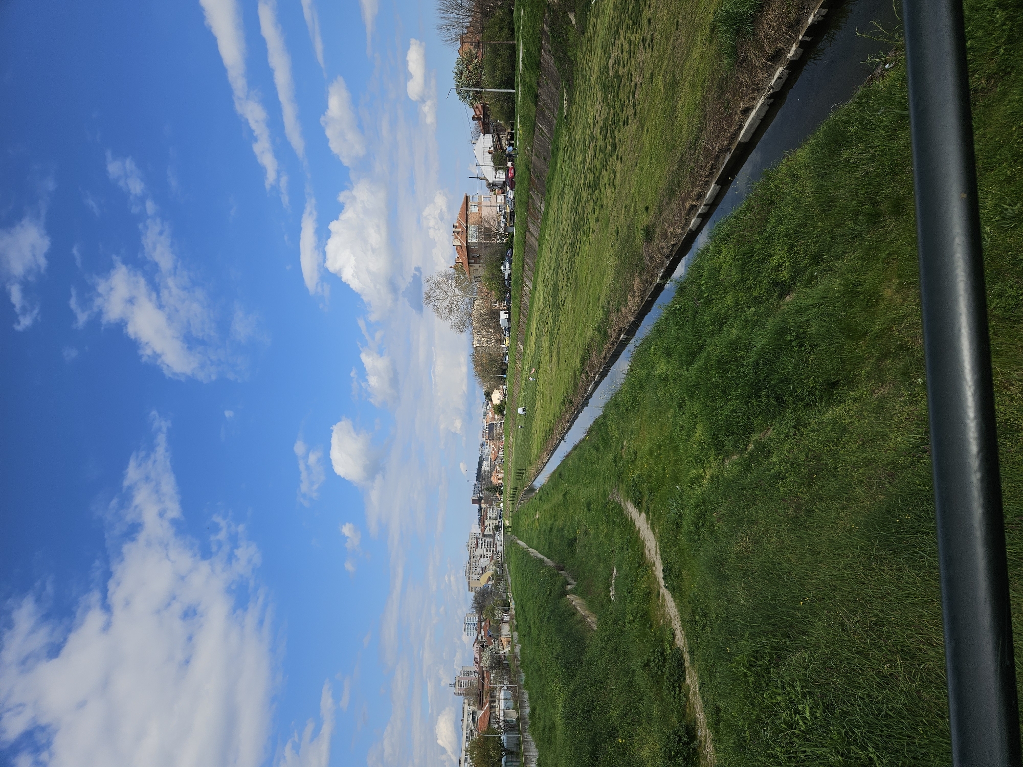 The image shows an elevated perspective of the Tundzha River in Yambol, Bulgaria, looking towards the city. The river flows through a concrete-lined channel flanked by grassy banks on both sides. The cityscape is visible in the distance, featuring buildings of varying heights and architectural styles. The sky is predominantly blue with scattered white clouds. A dark gray vertical element, possibly a railing or pole, is visible in the right side of the image.