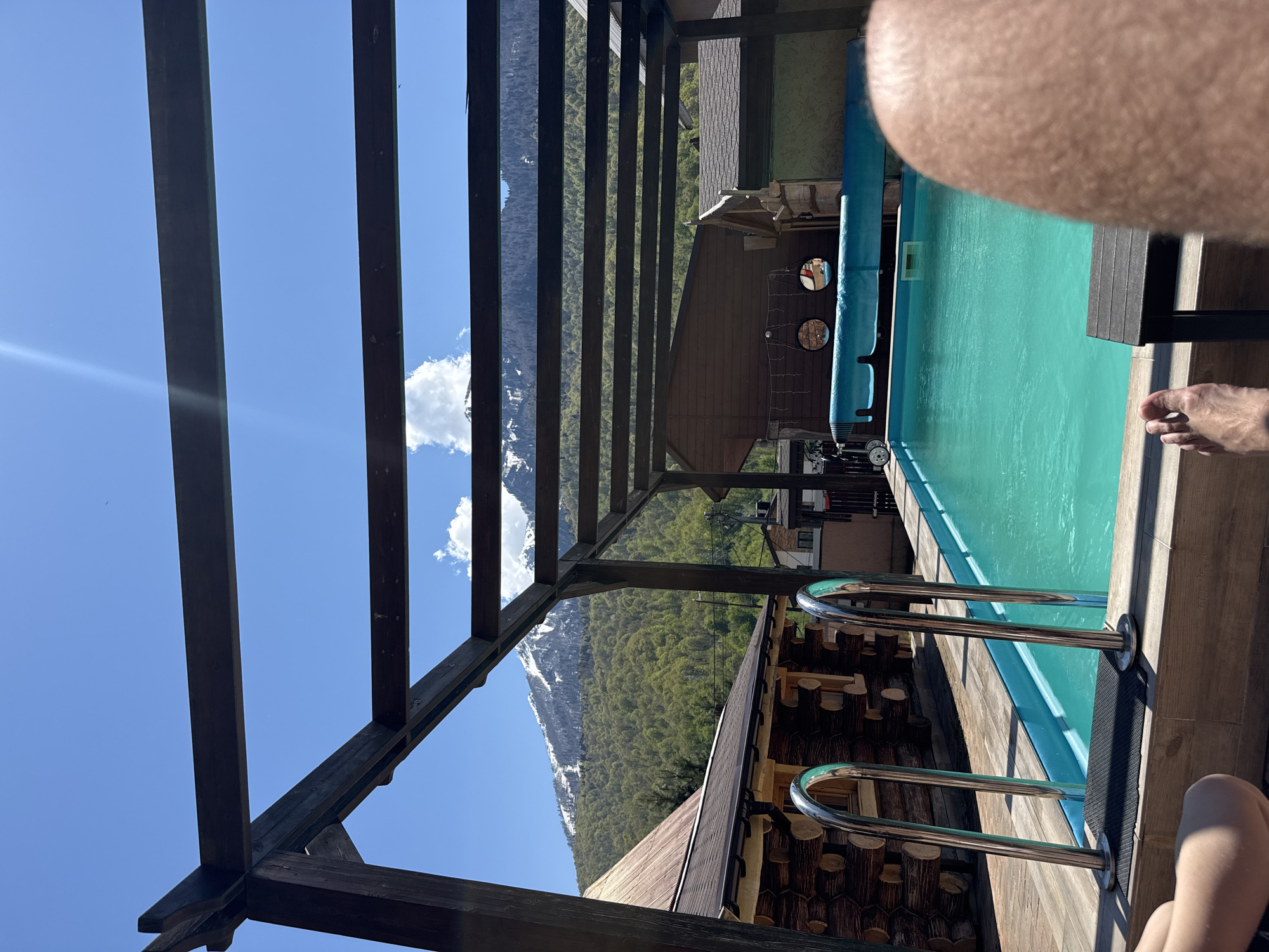 The image shows a partially obstructed view of a swimming pool framed by a wooden pergola and the surrounding landscape. The swimming pool is turquoise and appears to be outdoors. To the left, visible through the pergola, are mountains covered in trees and snow. The sky is blue with a few clouds. The pergola is made of dark wood. In the foreground, parts of a person's body are visible. The scene is likely located in a mountainous region, consistent with the Krasnaya Polyana area in Russia.