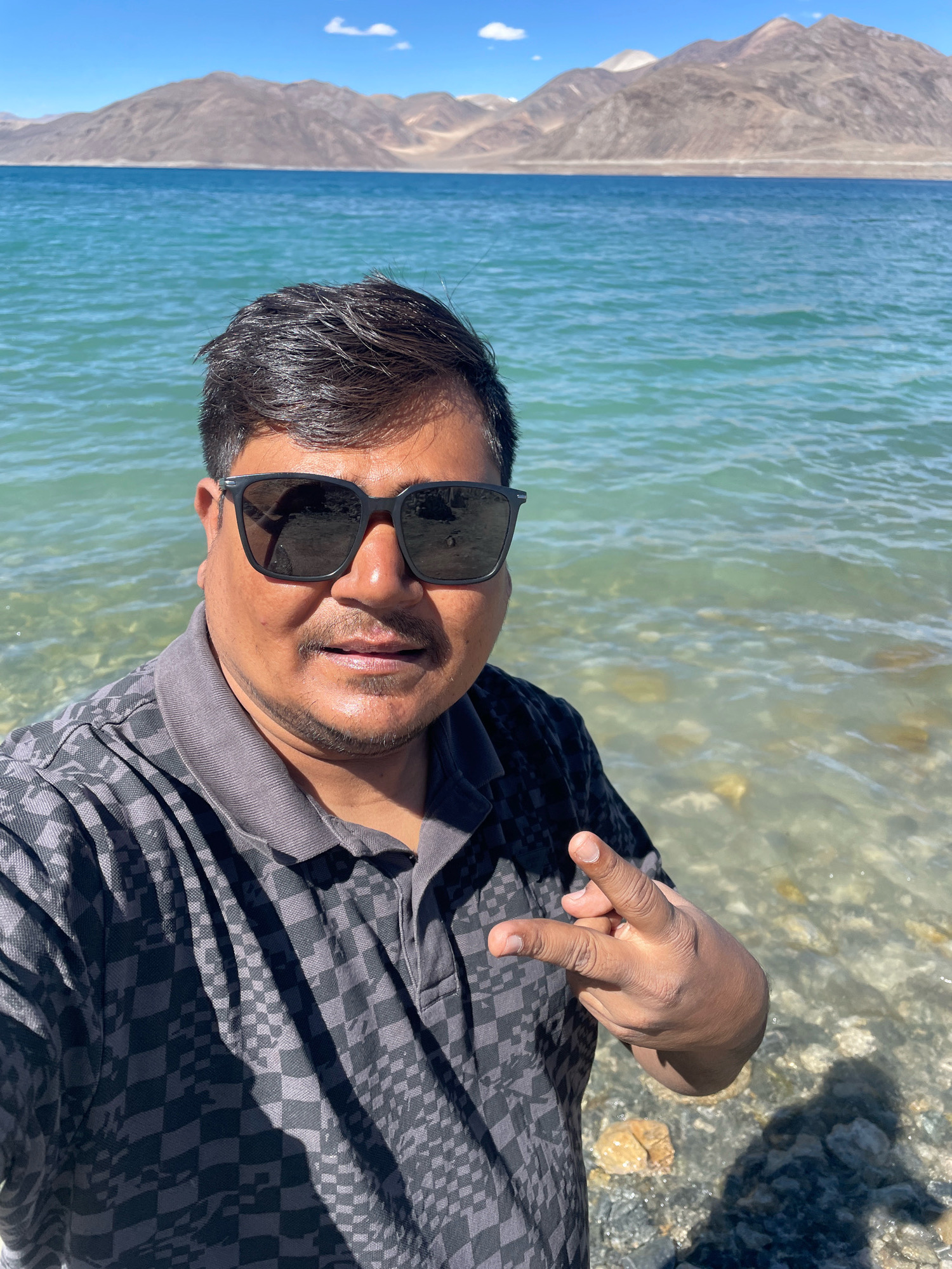 A man in a dark checkered polo shirt and sunglasses is taking a selfie outdoors. He is standing near the rocky shore of a clear blue lake, with a mountain range in the background. The man is making a peace sign with his right hand. The scene appears to be set in a natural, mountainous environment, possibly a lake in the Himalayas, given the context of "Man, India" as a potential location (though Man is a city in Ivory Coast, not India). The sky is blue with a few white clouds. The water is calm with some ripples.